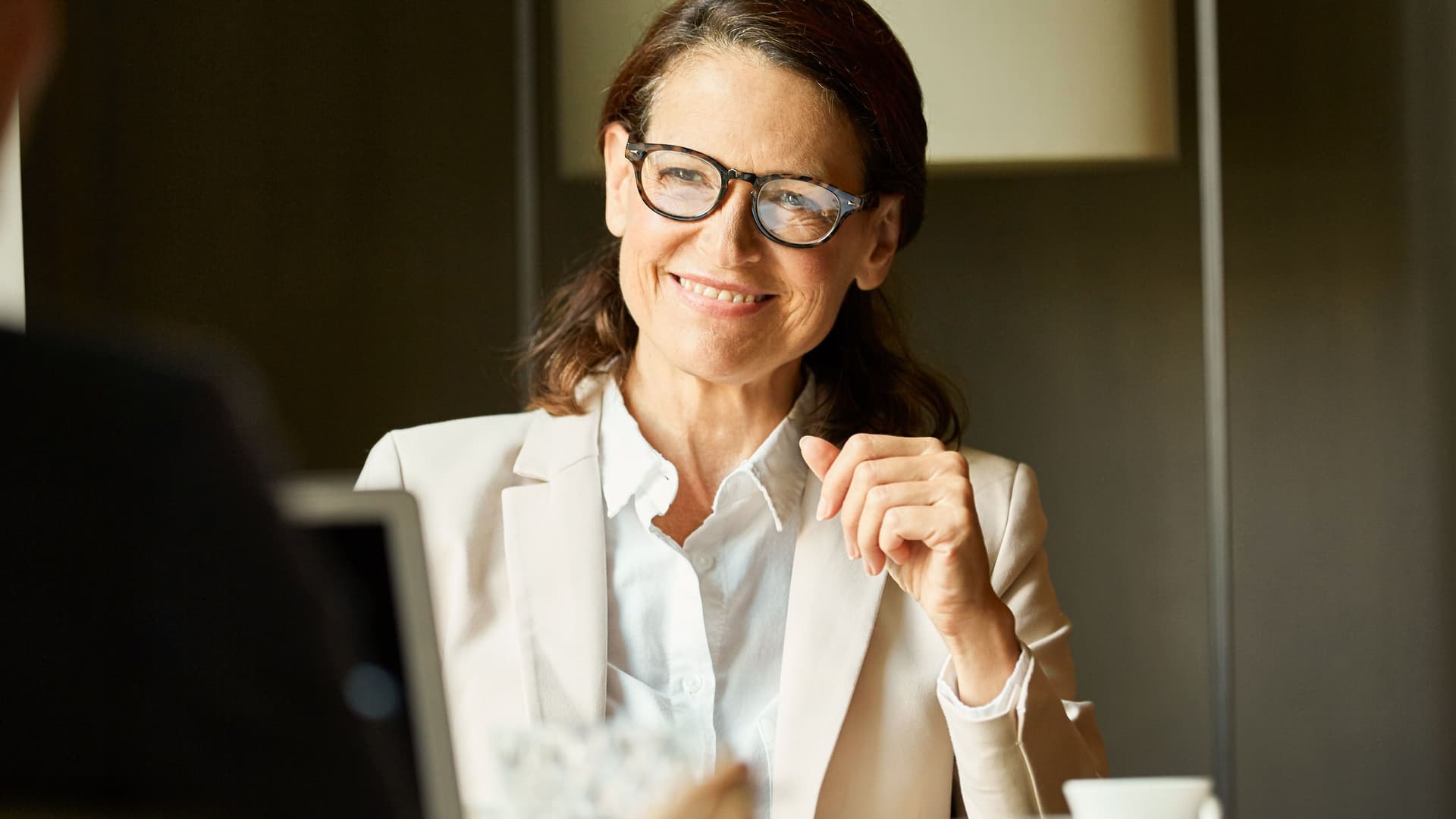 Frau in Business-Kleidung blickt lächelnd während einer Besprechung an einem Tisch zu ihrem Gegenüber.