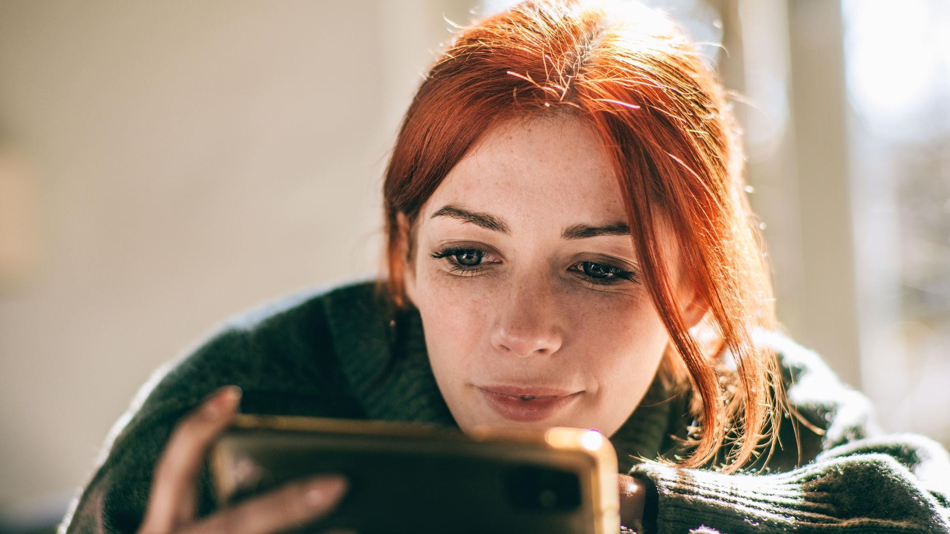 Close-up einer junge Frau mit roten Haaren, die auf ihr Mobiltelefon vor sich schaut.