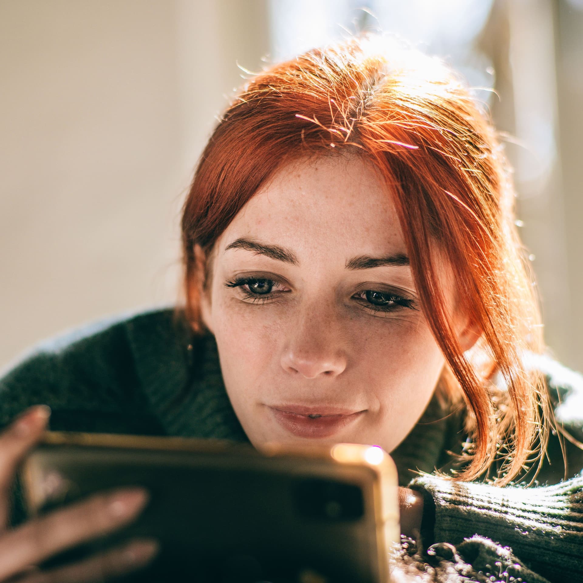 Close-up einer junge Frau mit roten Haaren, die auf ihr Mobiltelefon vor sich schaut.
