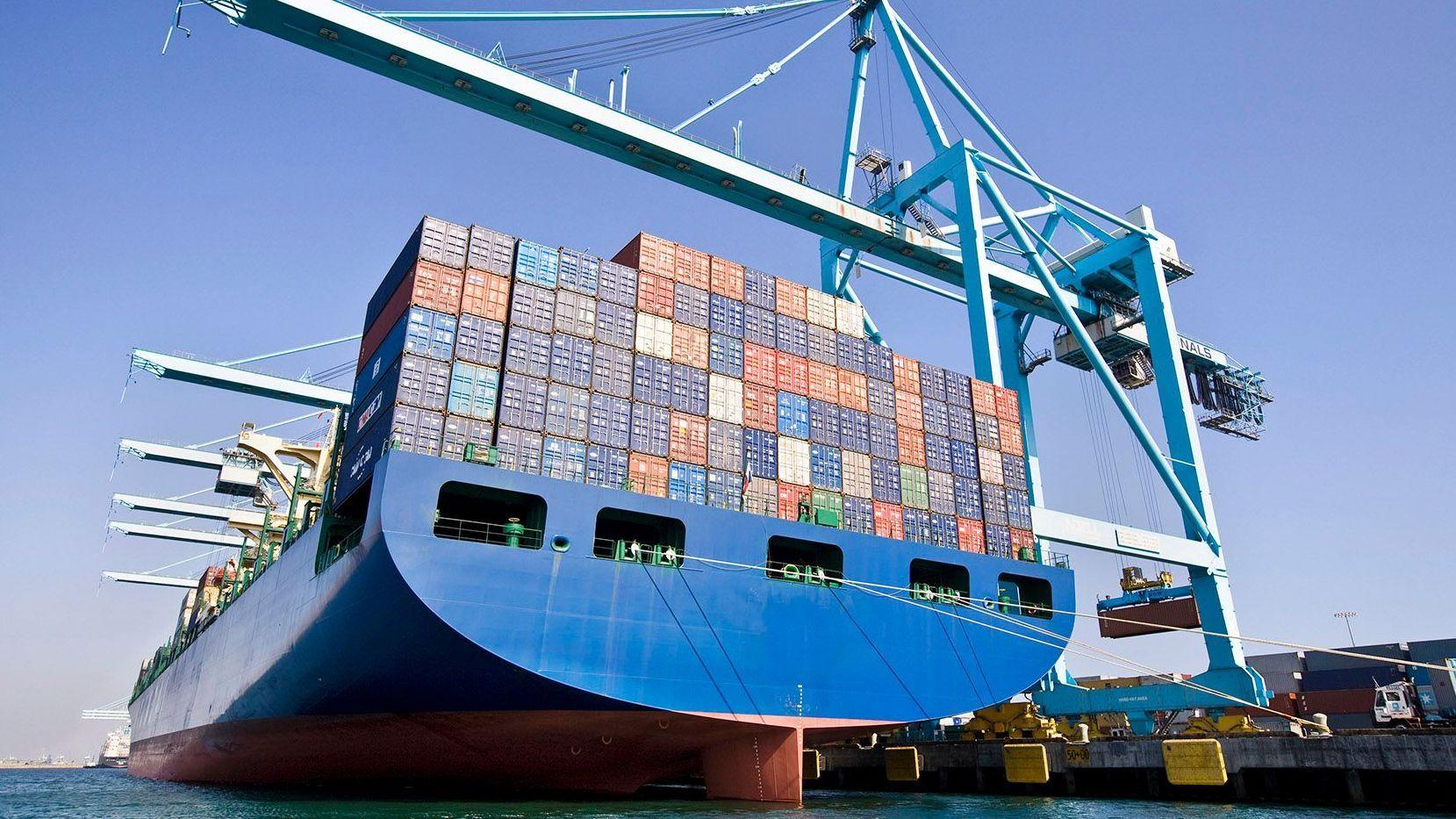 Ein großes Containerschiff liegt an einem Hafen. Über den bunten, gestapelten Containern ist ein Kran vor blauem Himmel zu sehen. 