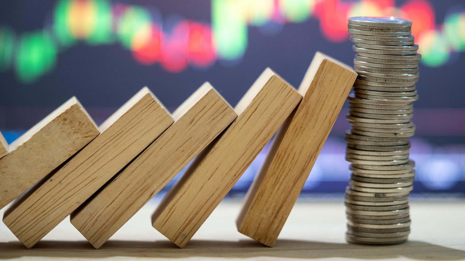Dominosteine lehnen gegen einen Stapel Münzen. Im Hintergrund sind Aktienkurse auf einem Bildschirm zu sehen.