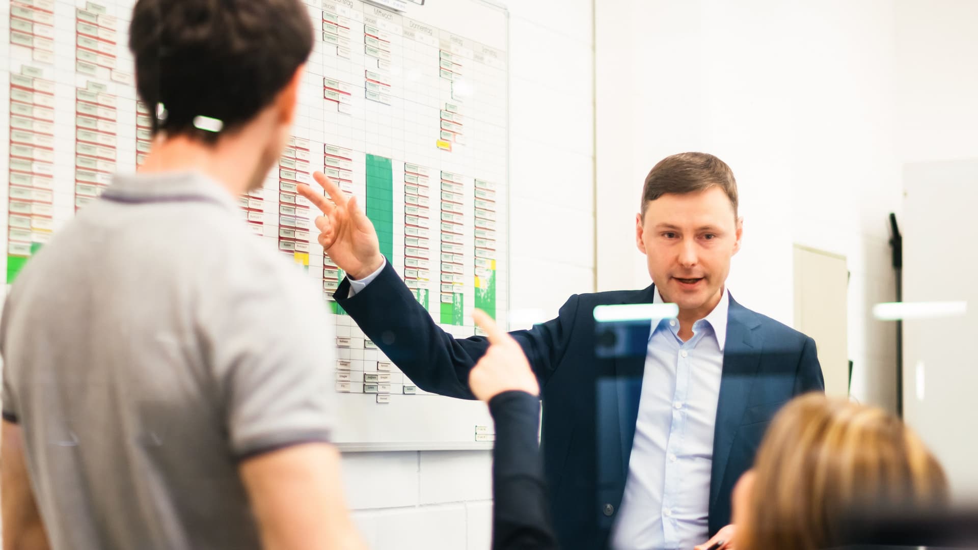 Zwei Männer und eine Frau bespreche etwas in einem hellen Industriebüro. Ein Mann und die Frau zeigt auf ein Whiteboard an der Wand.