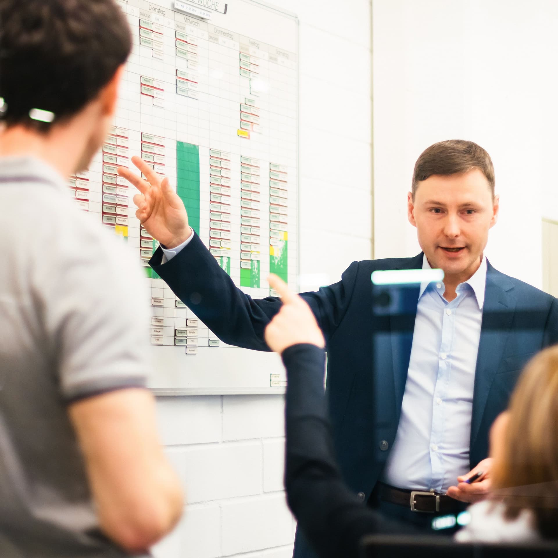 Zwei Männer und eine Frau bespreche etwas in einem hellen Industriebüro. Ein Mann und die Frau zeigt auf ein Whiteboard an der Wand.