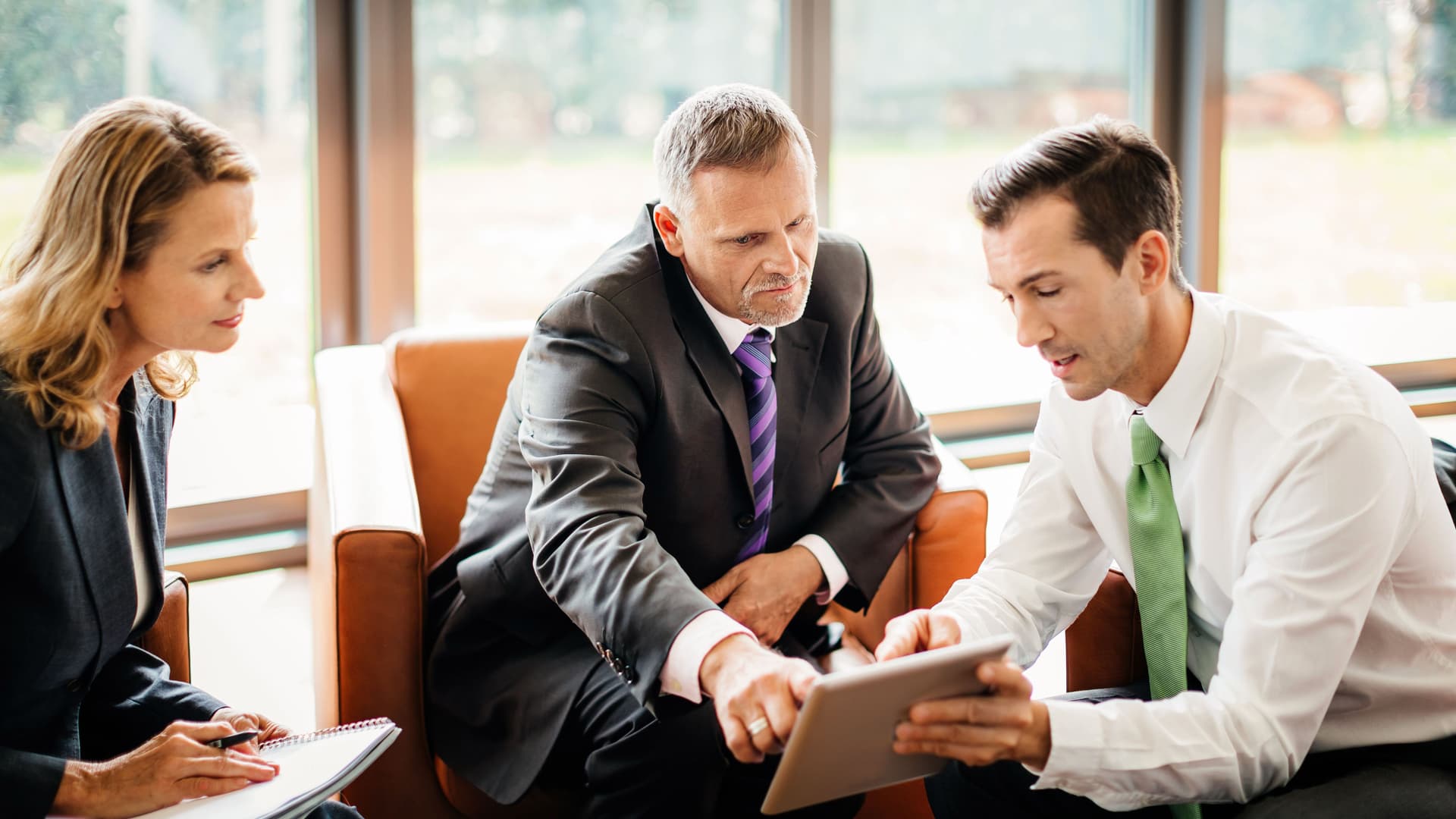 Zwei Männer und eine Frau in Business-Kleidung sitzen zusammen. Sie blicken auf ein Tablet, das der eine in den Händen hält.