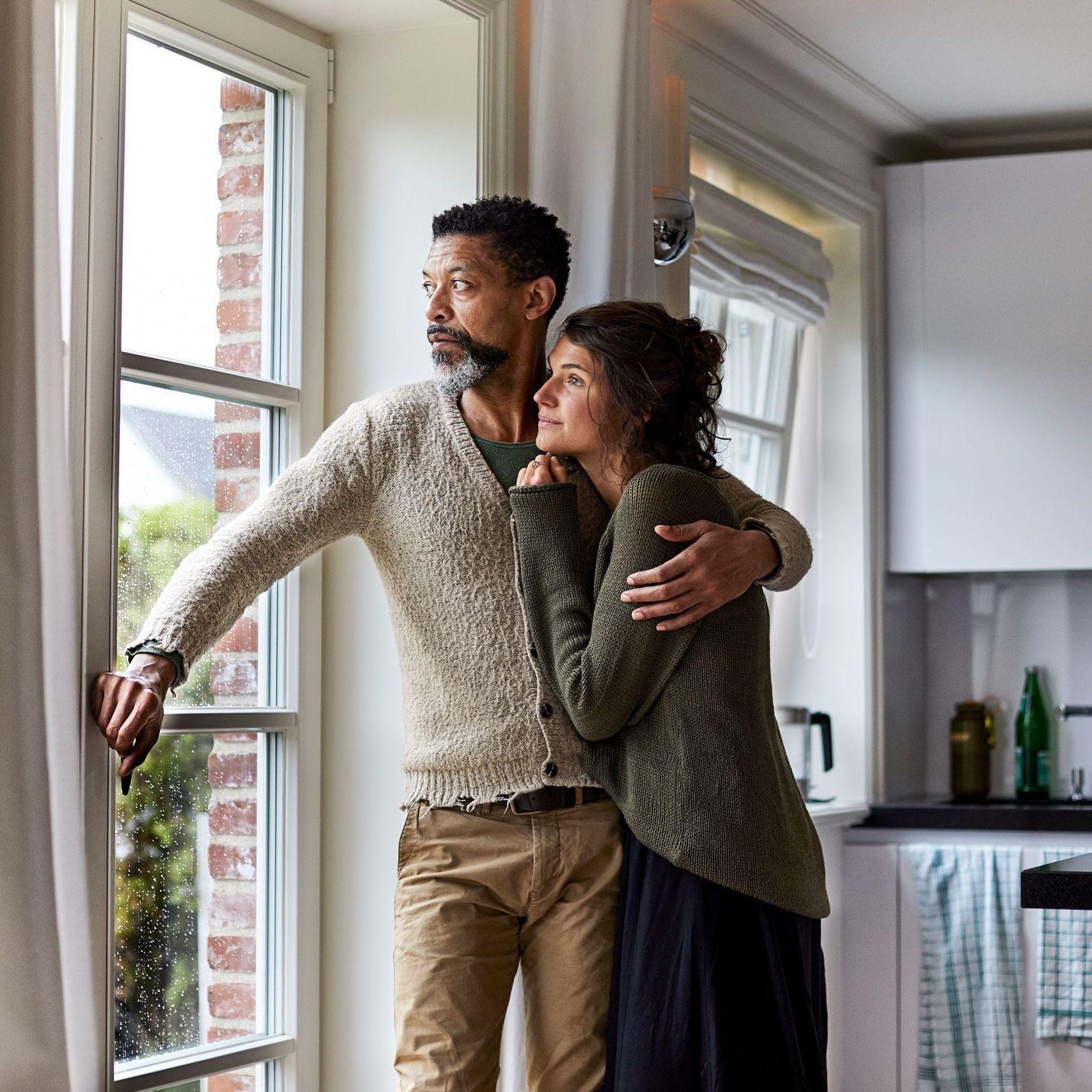 Ein sich umarmendes Paar mittleren Alters sieht zuhause nachdenklich, ernsthaft aus dem bodentiefen Fenster der Wohnküche.