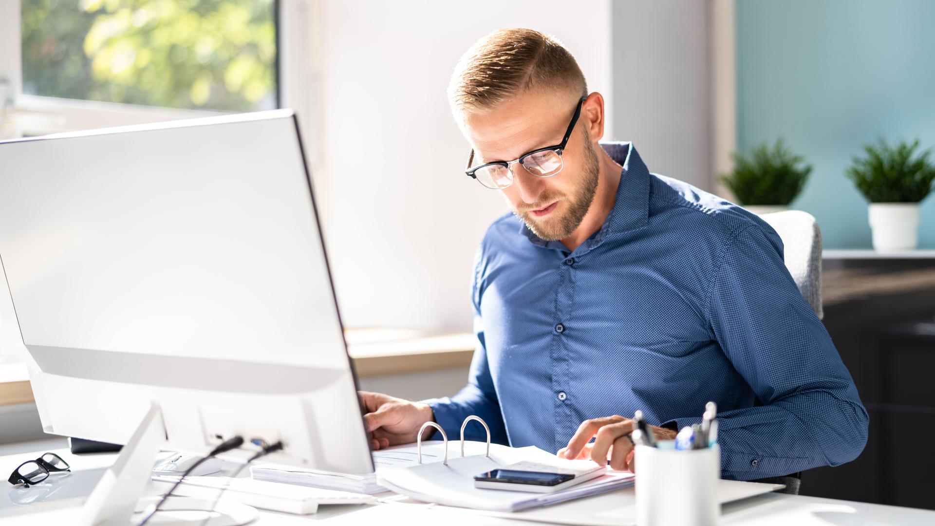 Ein Buchhalter im blauen Hemd mit Brille sitzt mit einem aufgeschlagenen Ordner und einem Taschenrechner vor seinem Computer.