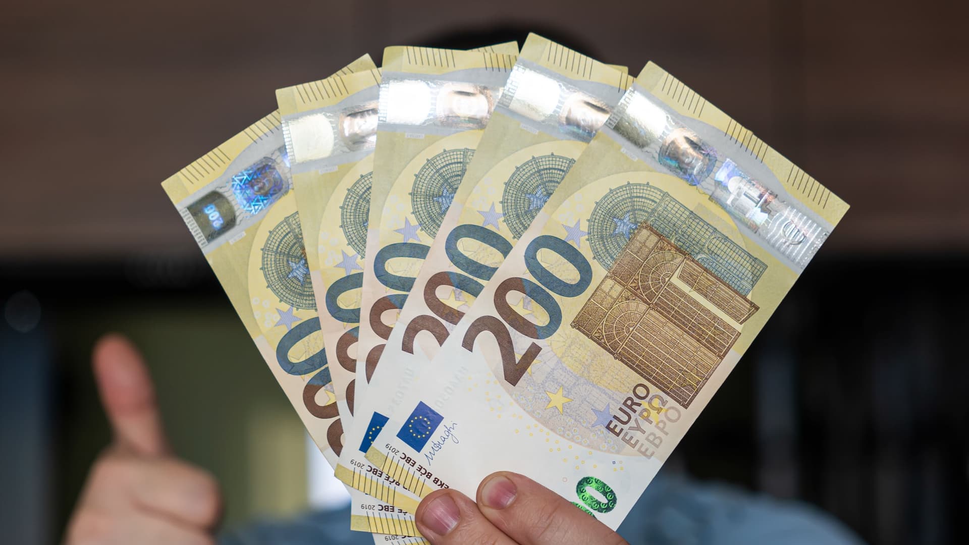 Eine Nahaufnahme zeigt eine Hand, die fünf 200-Euro-Banknoten fächerförmig hält. Die Banknoten sind goldfarben und zeigen typische Euro-Sicherheitsmerkmale. Im Hintergrund ist leicht unscharf ein Teil eines Daumens zu erkennen, der nach oben zeigt.