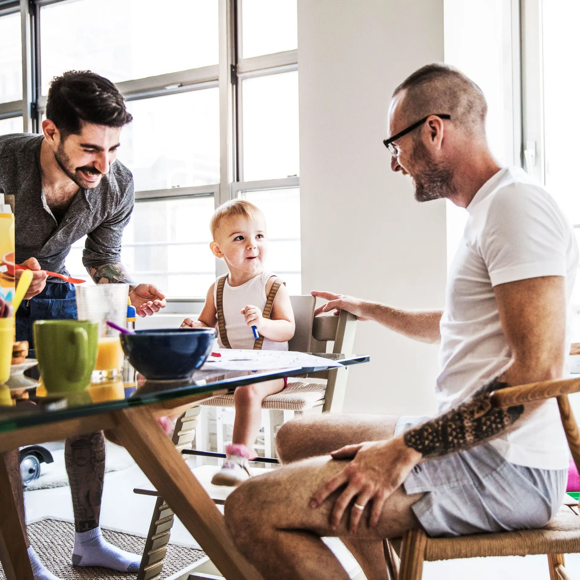 Ein homosexuelles männliches Paar sitzt mit seinem Kleinkind beim Frühstück. Alle lachen.