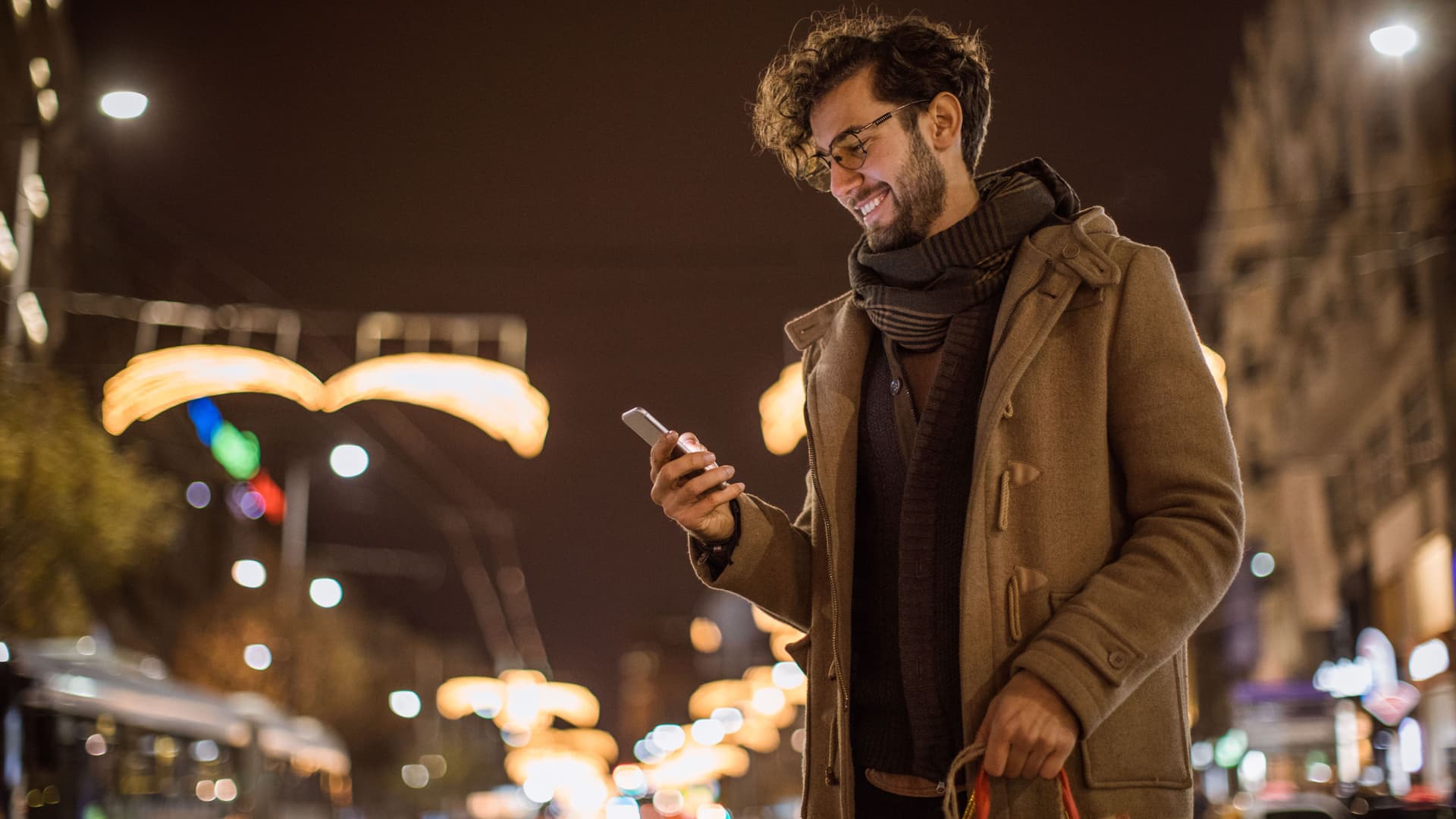 Ein Mann mit Einkaufstüten in der Hand schaut glücklich auf sein Smartphone. Im Hintergrund ist ein weihnachtlich erleuchteter Nachthimmel.