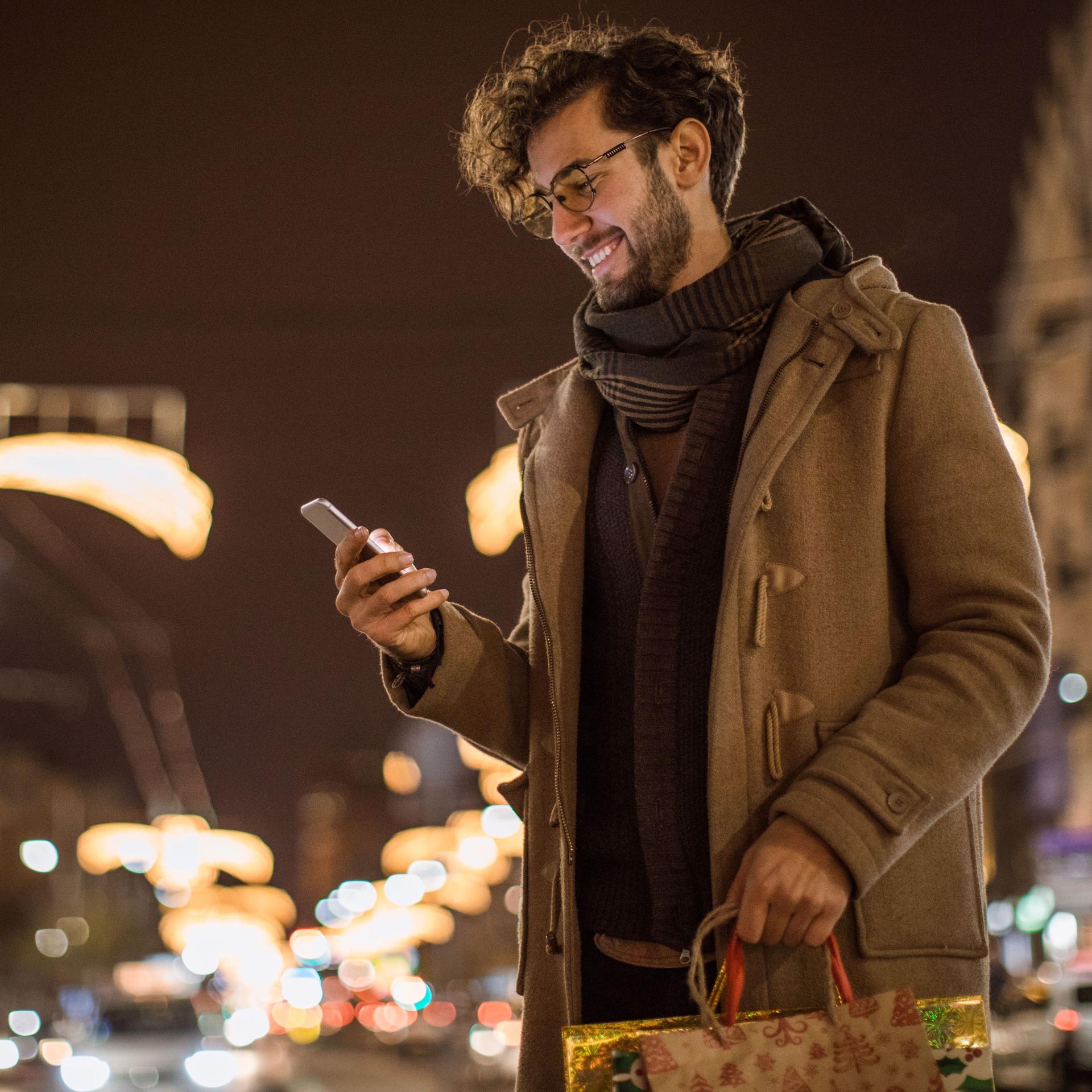 Ein Mann mit Einkaufstüten in der Hand schaut glücklich auf sein Smartphone. Im Hintergrund ist ein weihnachtlich erleuchteter Nachthimmel.