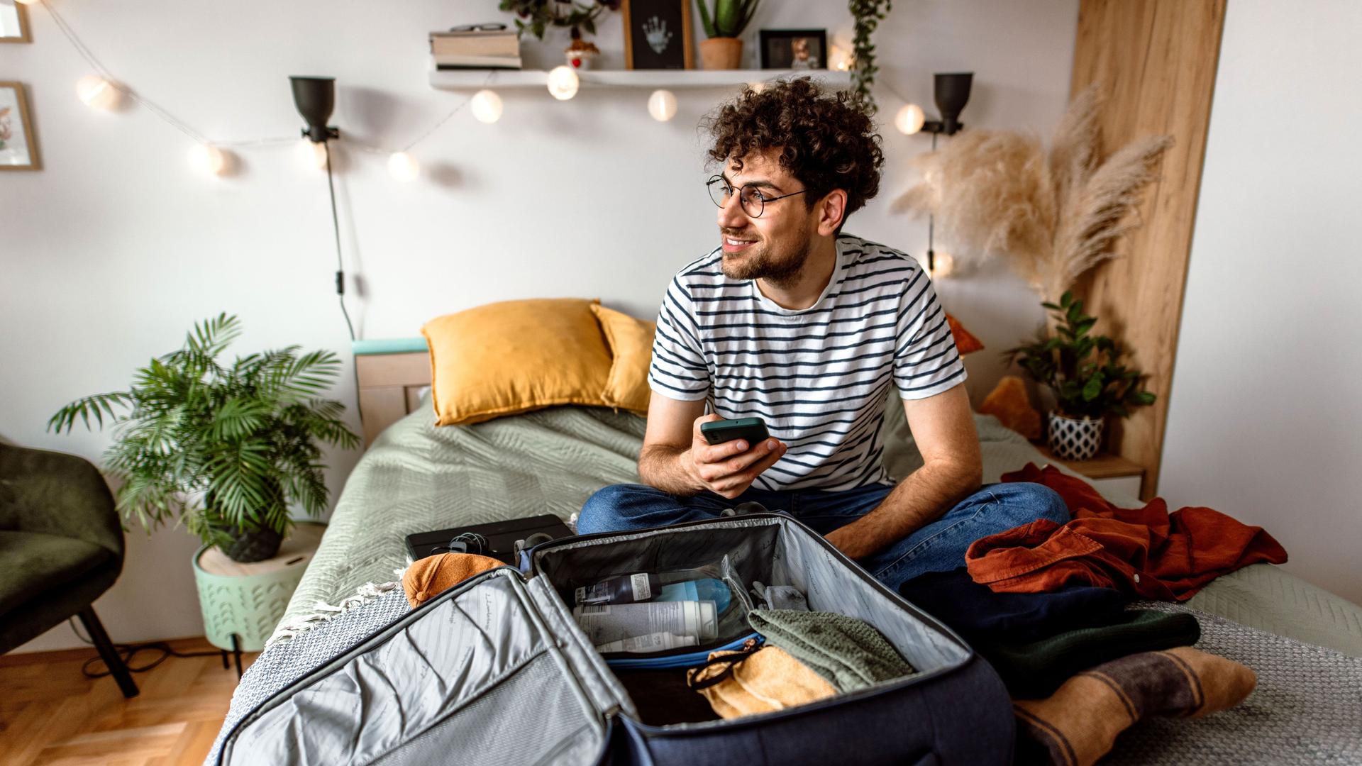 Ein junger Mann sitzt mit seinem Smartphone auf seinem Bett. Vor ihm liegt ein geöffneter Koffer.