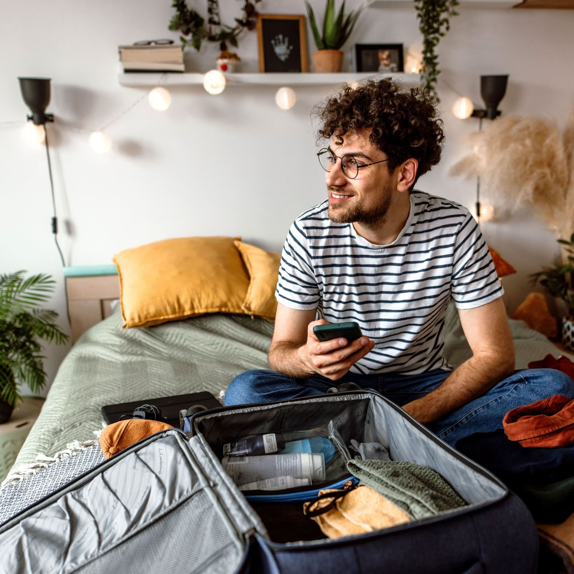 Ein junger Mann sitzt mit seinem Smartphone auf seinem Bett. Vor ihm liegt ein geöffneter Koffer.