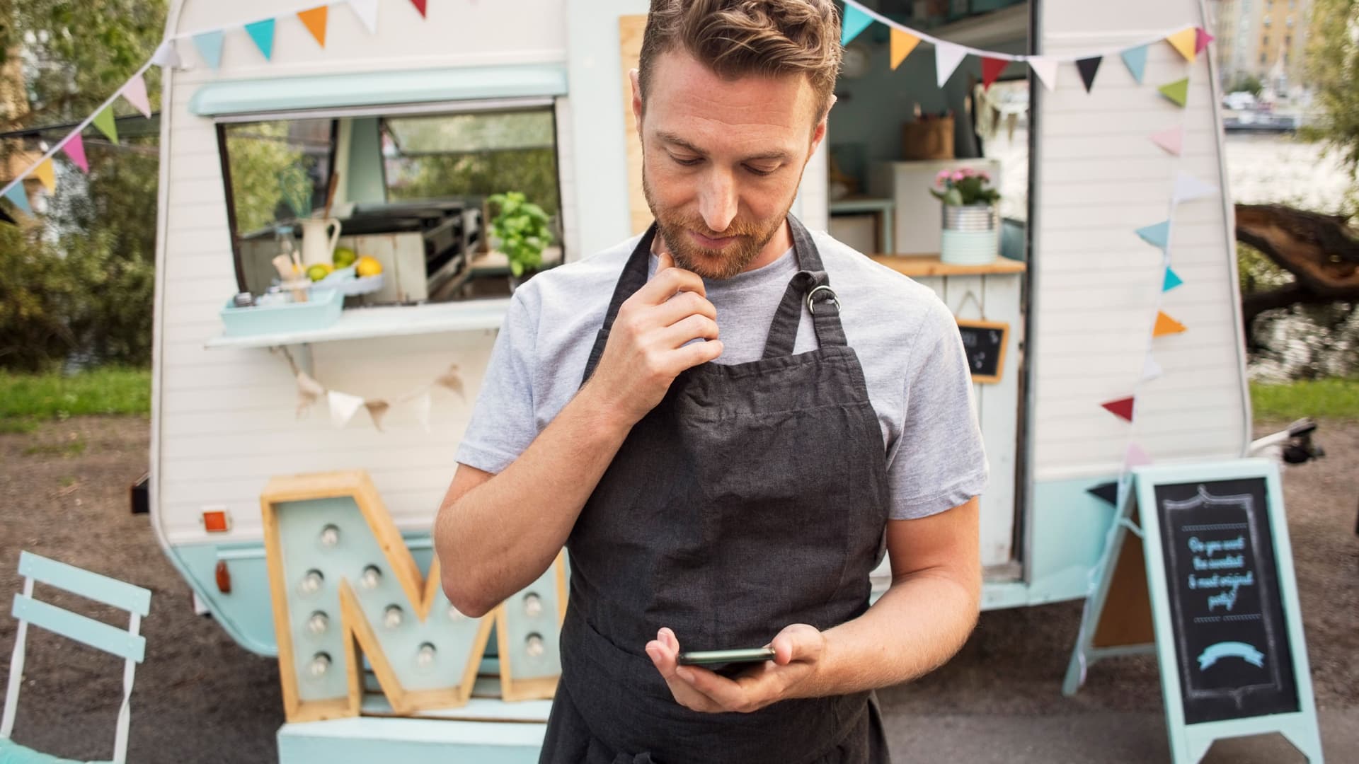 Ein Mann mit kurzen Haaren und schwarzer Schürze umgebunden schaut auf sein Smartphone in der Hand. Er steht vor seinem Imbisswagen.