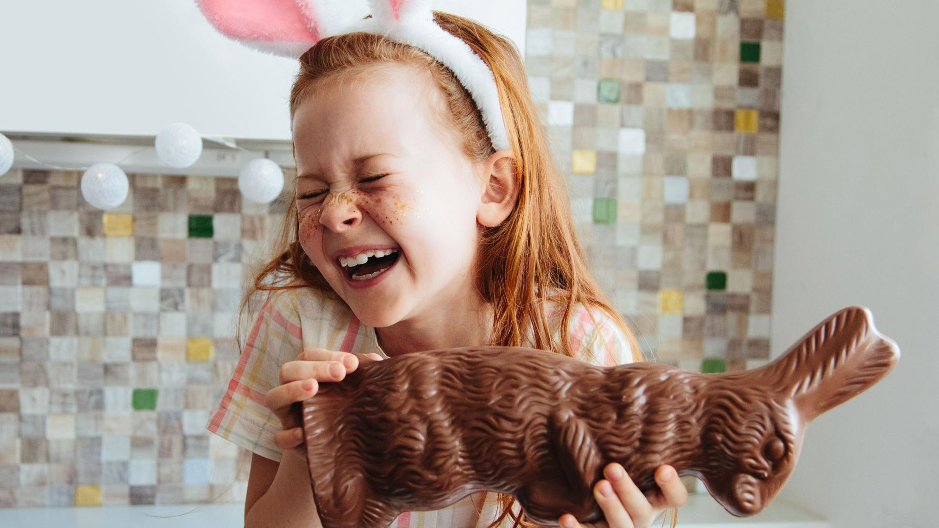 Ein Mädchen mit einem Hasenohr-Haarreif sitz mit einem sehr großen Schoko-Hassen in der Küche. Sie lacht.