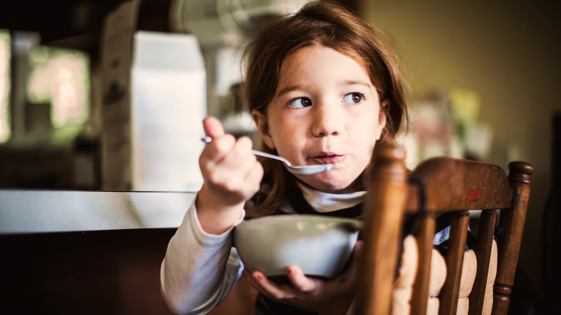 ein Mädchen sitzt mit einer Schüssel Müsli auf einem Stuhl. Sie hat einen Löffel in der Hand und dreht sich zur Kamera.