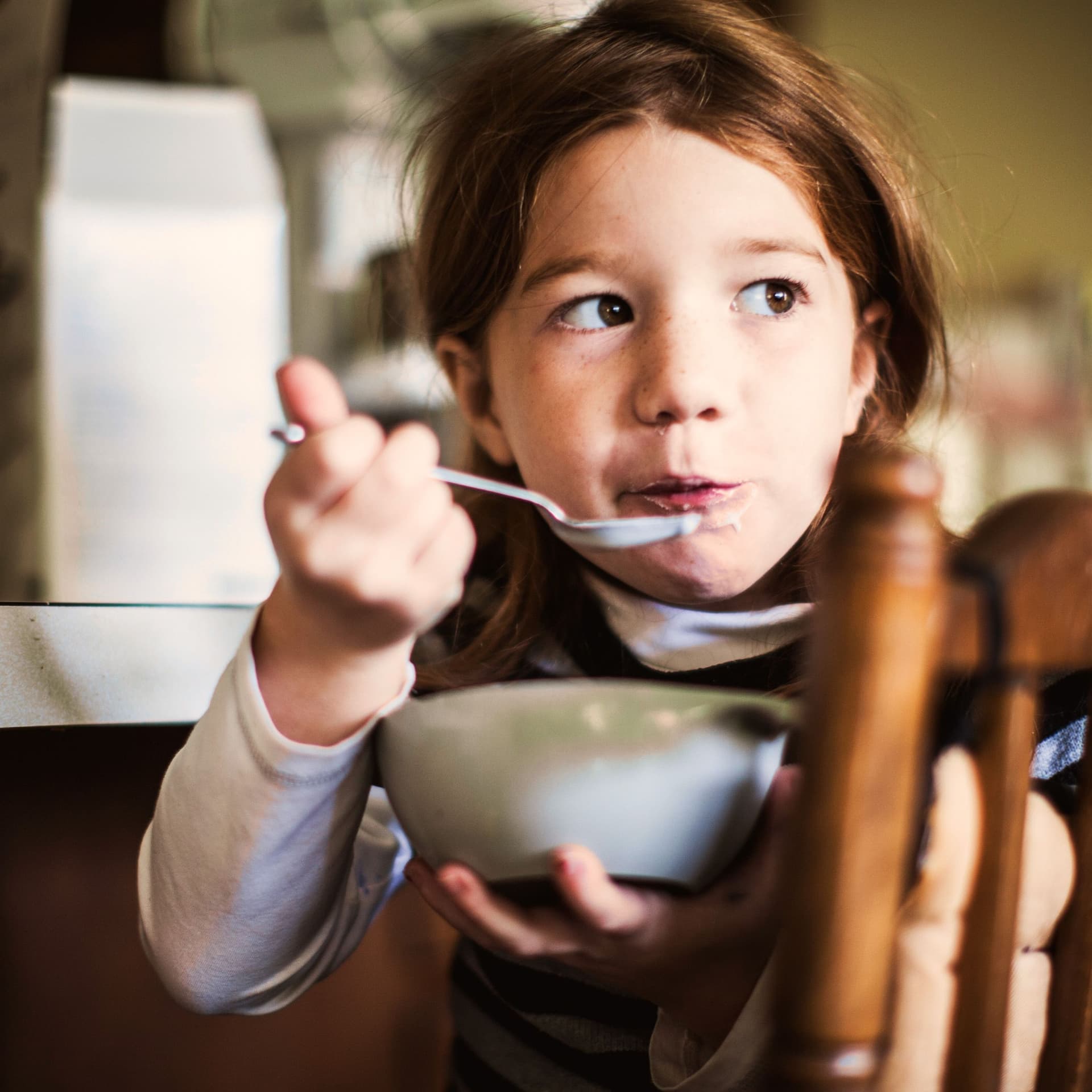 ein Mädchen sitzt mit einer Schüssel Müsli auf einem Stuhl. Sie hat einen Löffel in der Hand und dreht sich zur Kamera.