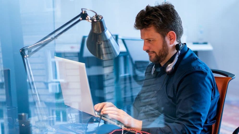 Ein Mann sitzt an einem Schribtisch vor einem Monitor. Der Blick ist durch eine Glasscheibe mit Spiegelungen.