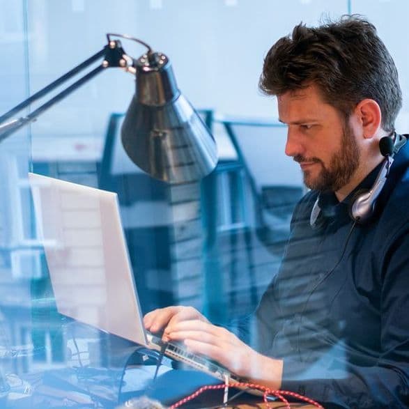 Ein Mann sitzt an einem Schribtisch vor einem Monitor. Der Blick ist durch eine Glasscheibe mit Spiegelungen.