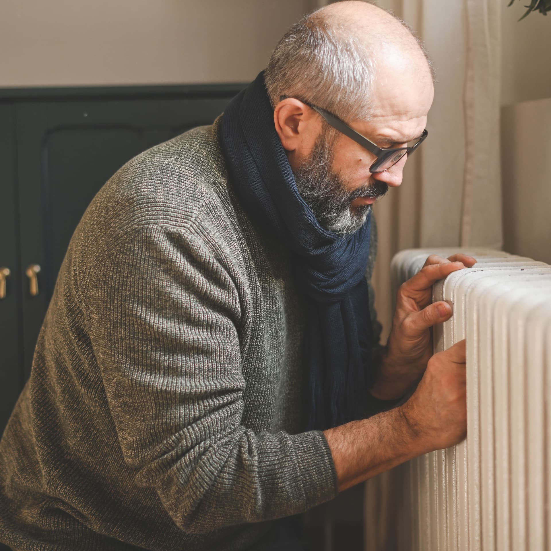 Ein Mann mit Wollpullover und Schal kniet vor einer Heizung.