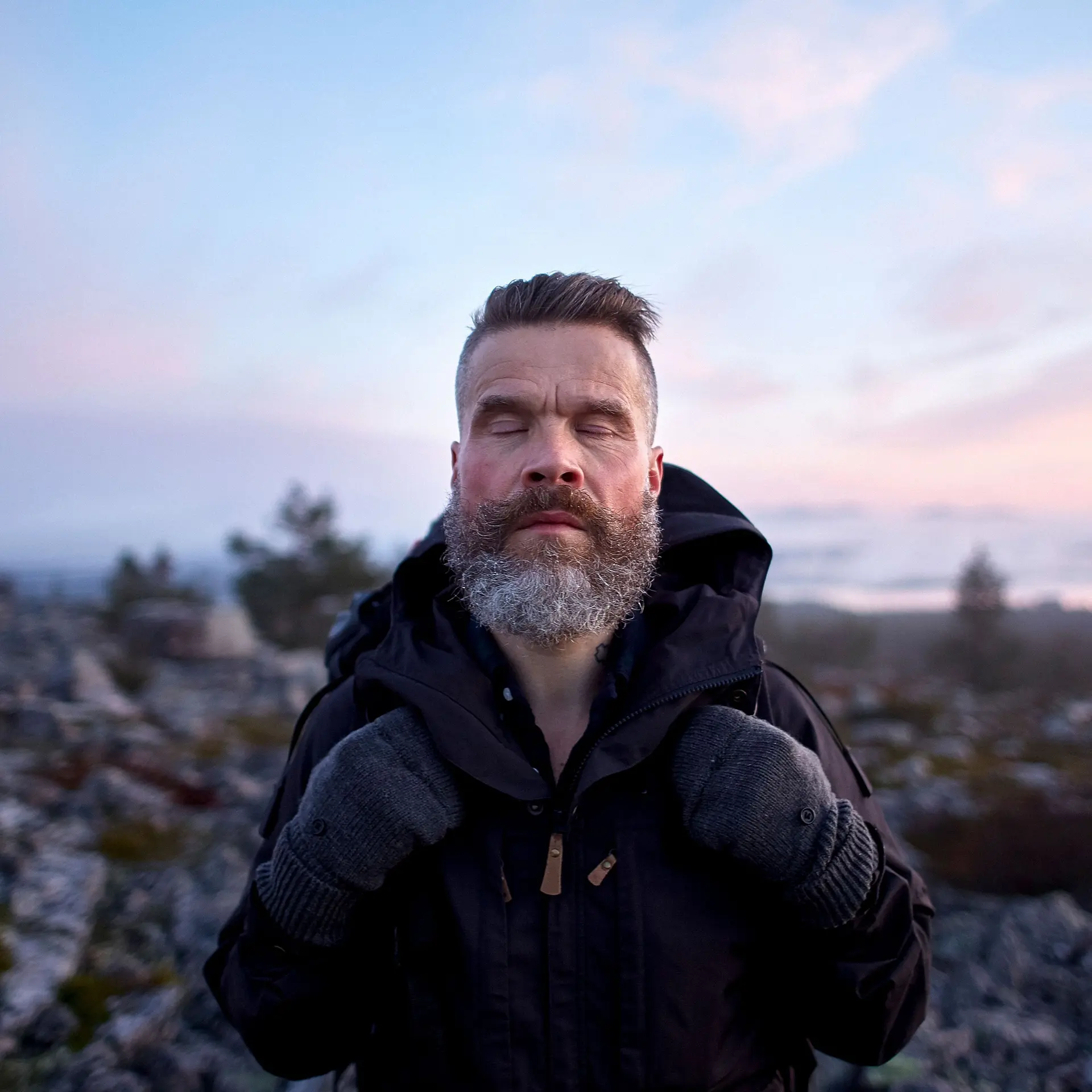 Ein Mann mit grauem Bart steht auf einem Berg mit geschlossenen Augen und genießt den Moment. DIe Sonne ist gerade untergegangen.