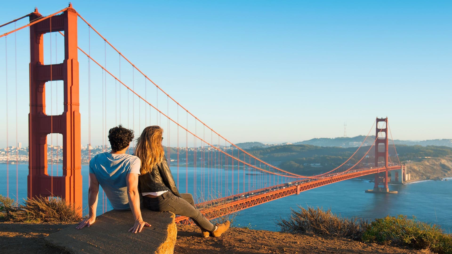 Ein Mann und eine Frau sitzen auf einem Stein und schauen auf die Golden Gate Bridge. Im Hintergrund ist San Francisco zu sehen.