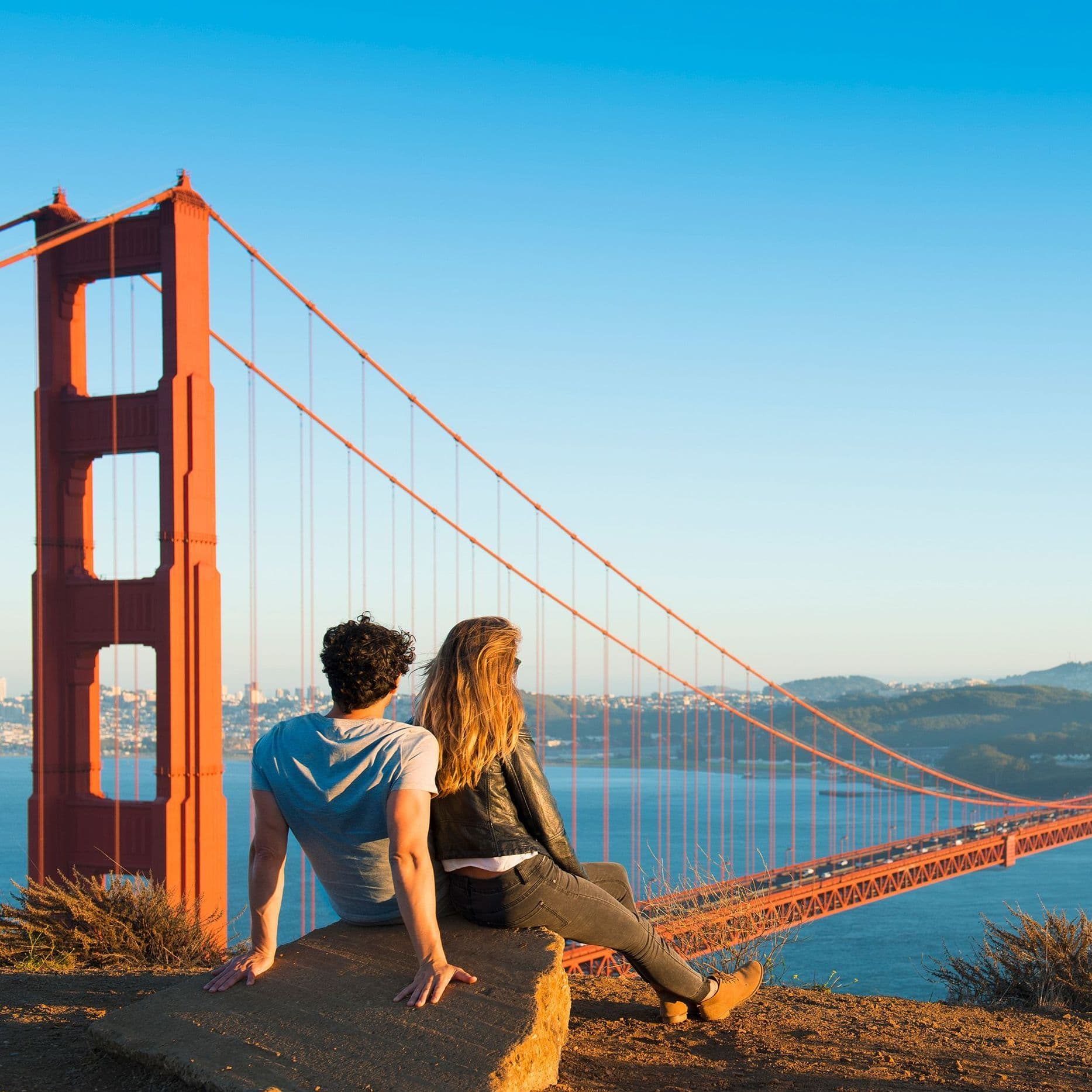 Ein Mann und eine Frau sitzen auf einem Stein und schauen auf die Golden Gate Bridge. Im Hintergrund ist San Francisco zu sehen.