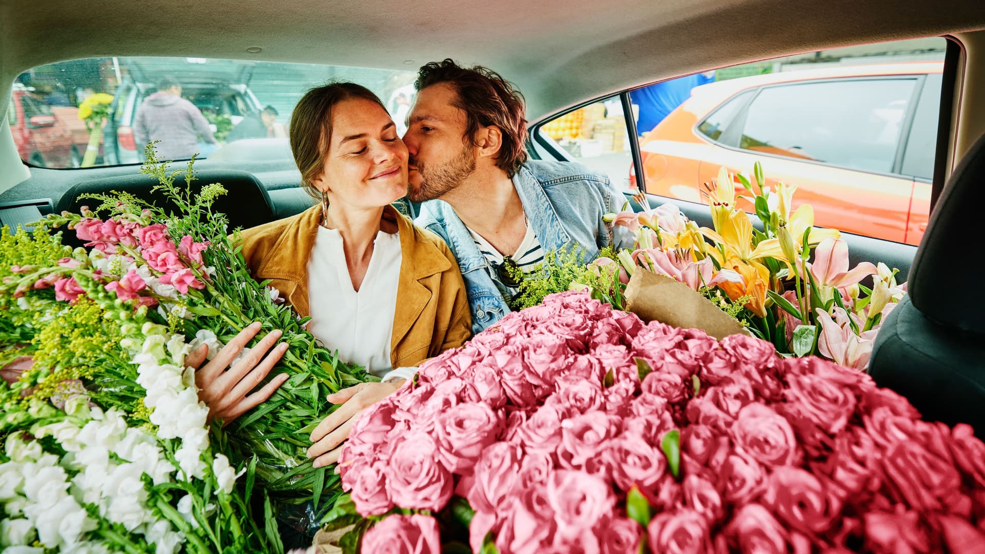 Ein Mann und eine Frau sitzen auf dem Rücksitz eines Autos. Das Auto ist voll mit bunten Schnittblumen.