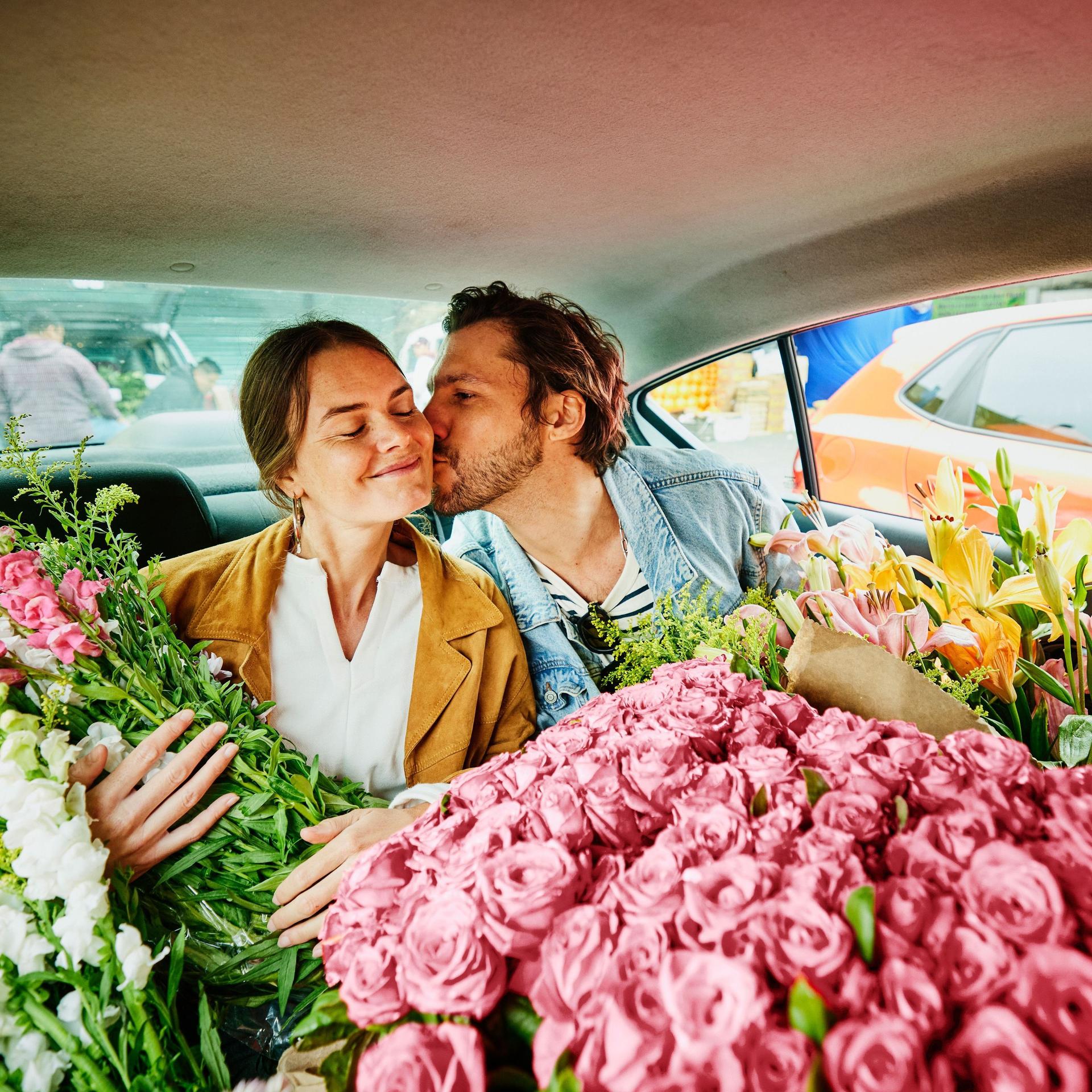 Ein Mann und eine Frau sitzen auf dem Rücksitz eines Autos. Das Auto ist voll mit bunten Schnittblumen.