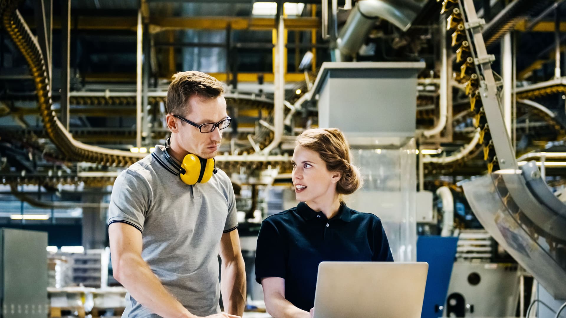 Ein Mann und eine Frau stehen vor einer Produktionsstraße. Sie schauen auf einen Laptop und unterhalten sich.