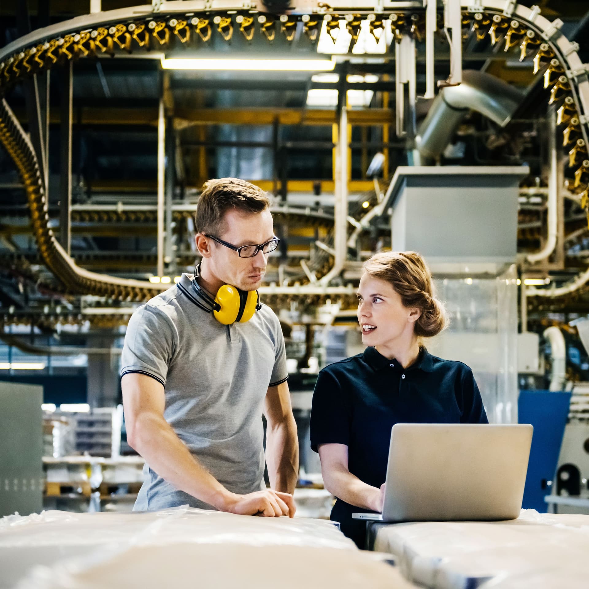 Ein Mann und eine Frau stehen vor einer Produktionsstraße. Sie schauen auf einen Laptop und unterhalten sich.