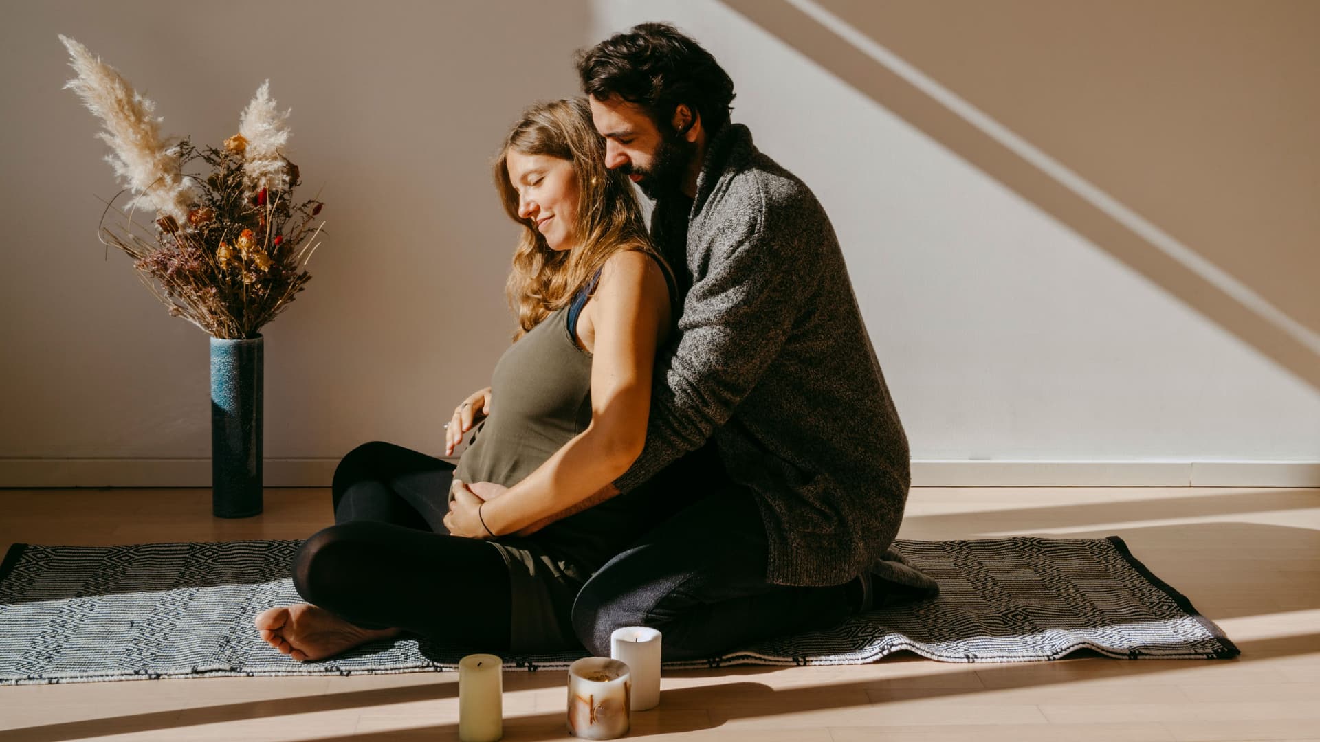 Ein Mann und eine Frau sitzen gemeinsam auf dem Boden in der Sonne. Die Frau ist schwanger. Der Mann umarmt sie von hinten.