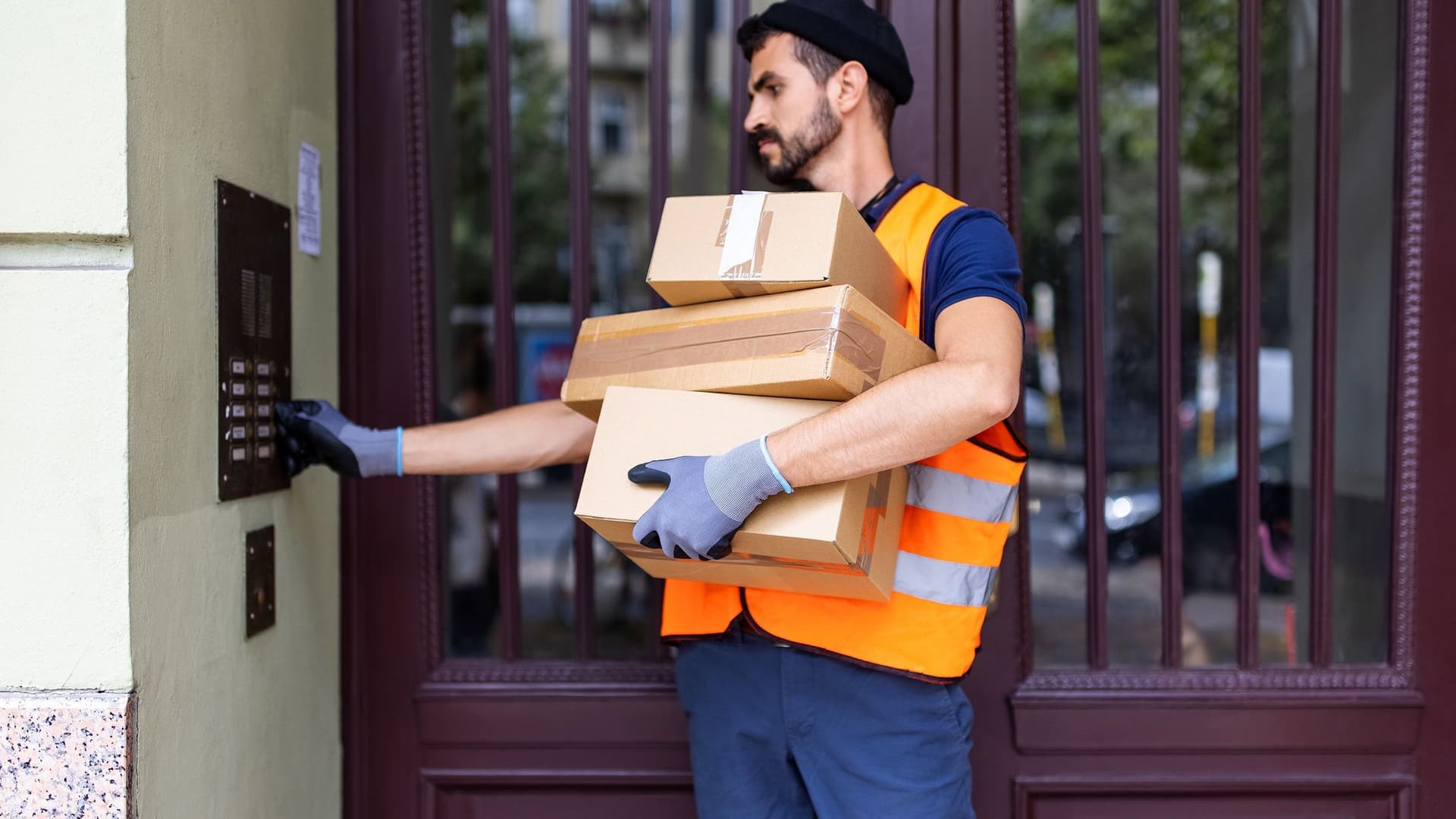 Ein Paketbote klingelt an einem Mehrfamilienhaus. Er hat den Arm voller Pakete.