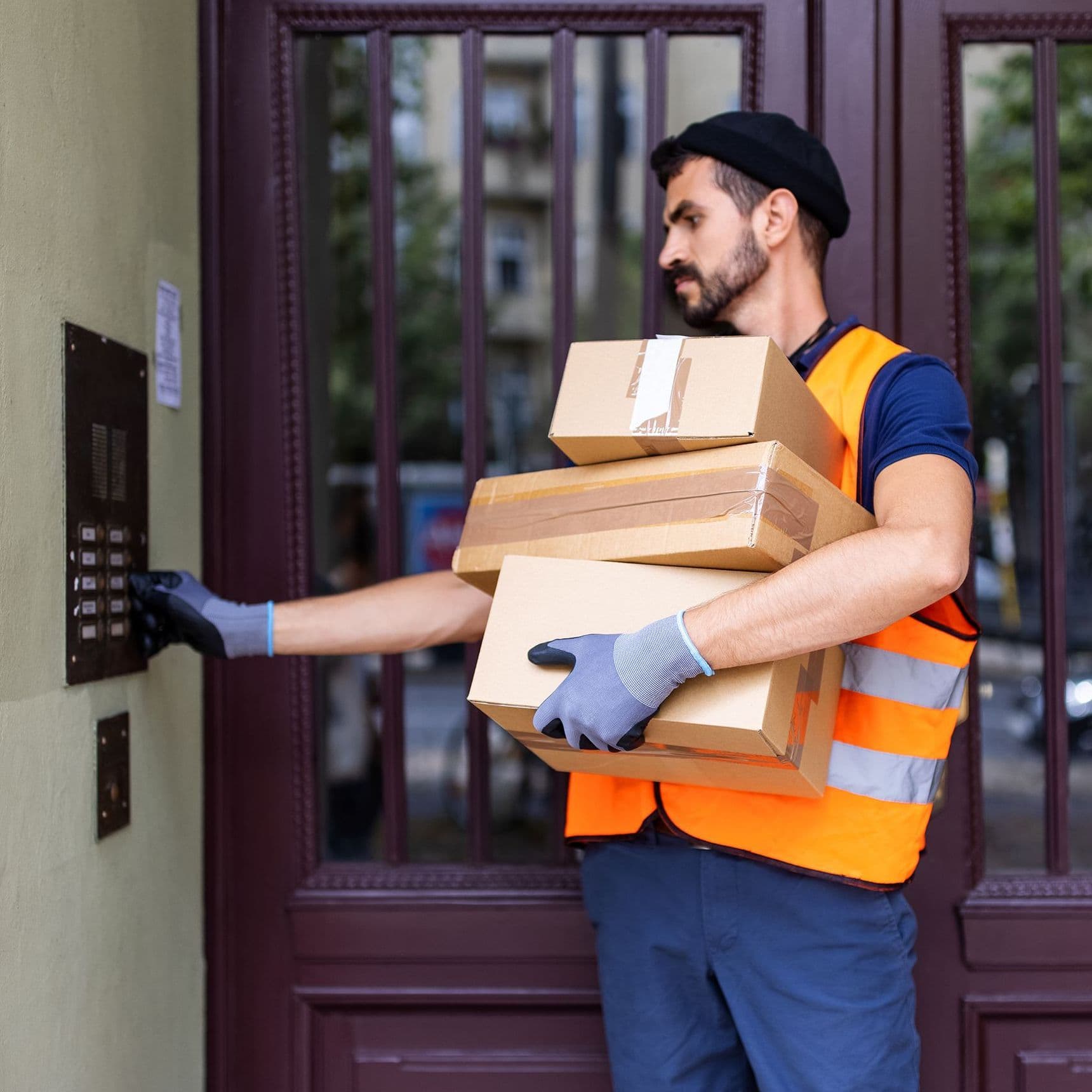 Ein Paketbote klingelt an einem Mehrfamilienhaus. Er hat den Arm voller Pakete.