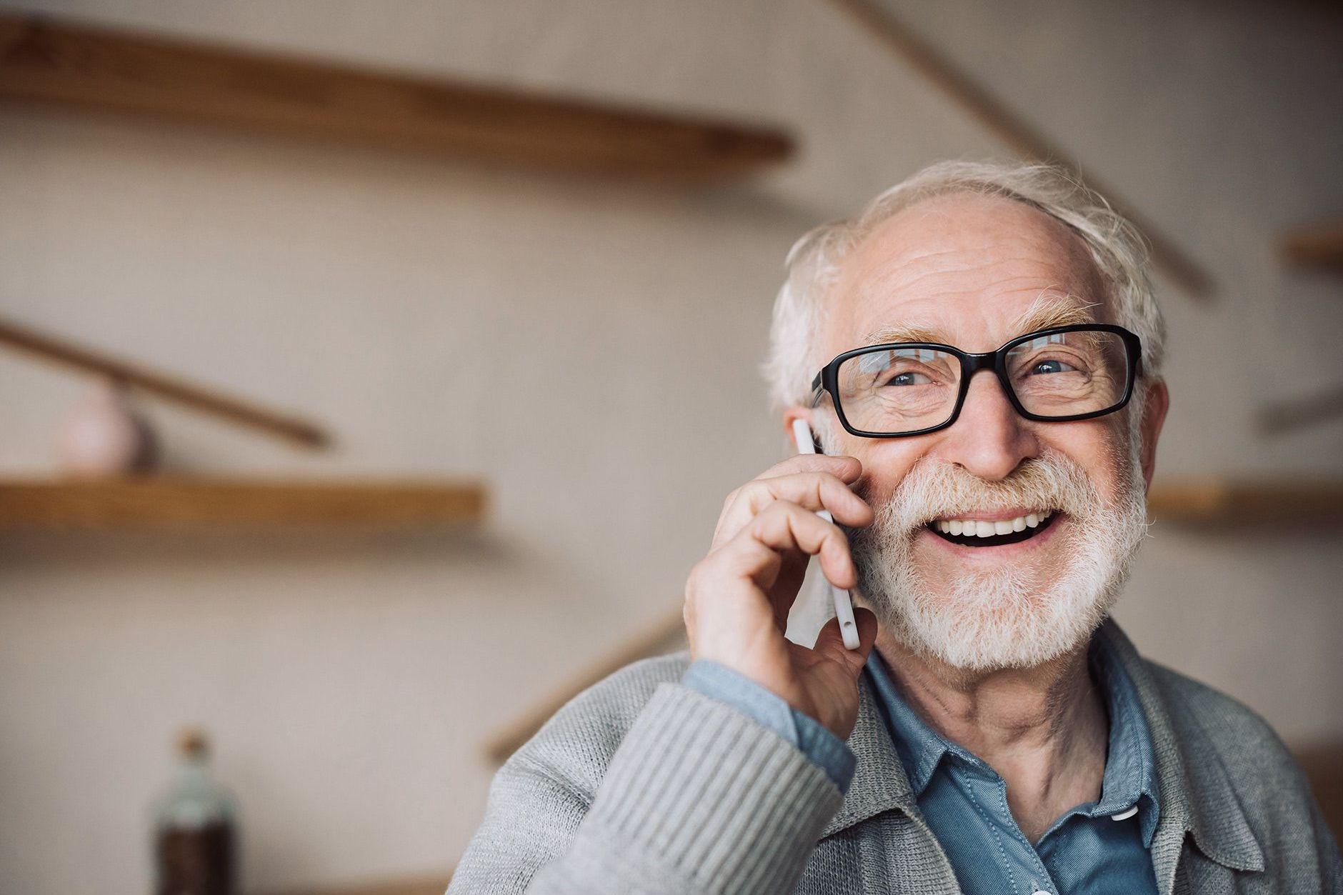 Ein Senior führt lächelnd ein Telefongespräch in seinem Wohnbereich und hat ein Smartphone am Ohr. 