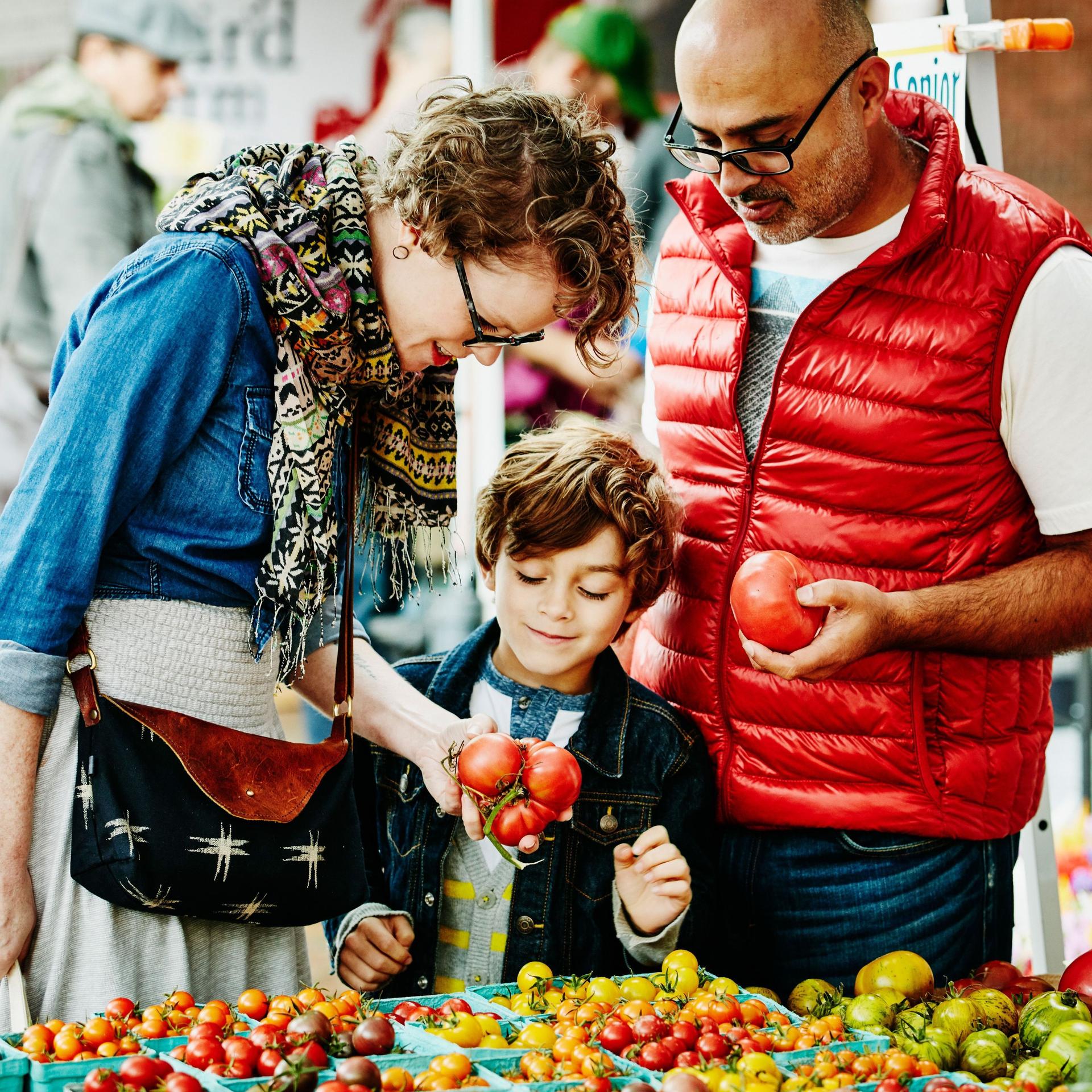 Eine Familie geht auf einem Wochenmarkt einkaufen. Sie schauen sich die Tomaten an.