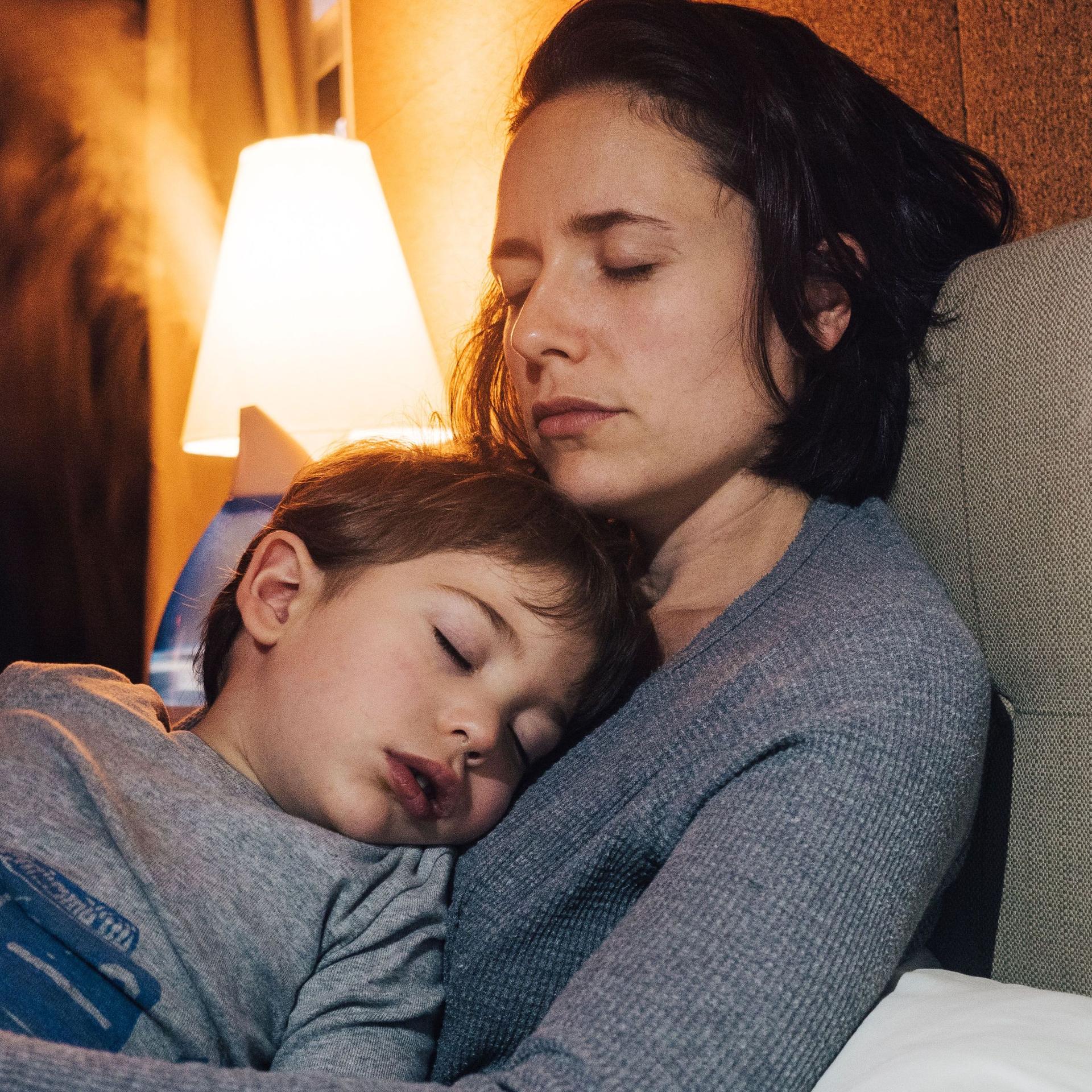 Eine Frau mit geschlossenen Augen sitzt auf dem Bett. Sie hält ihr krankes Kind im Arm.