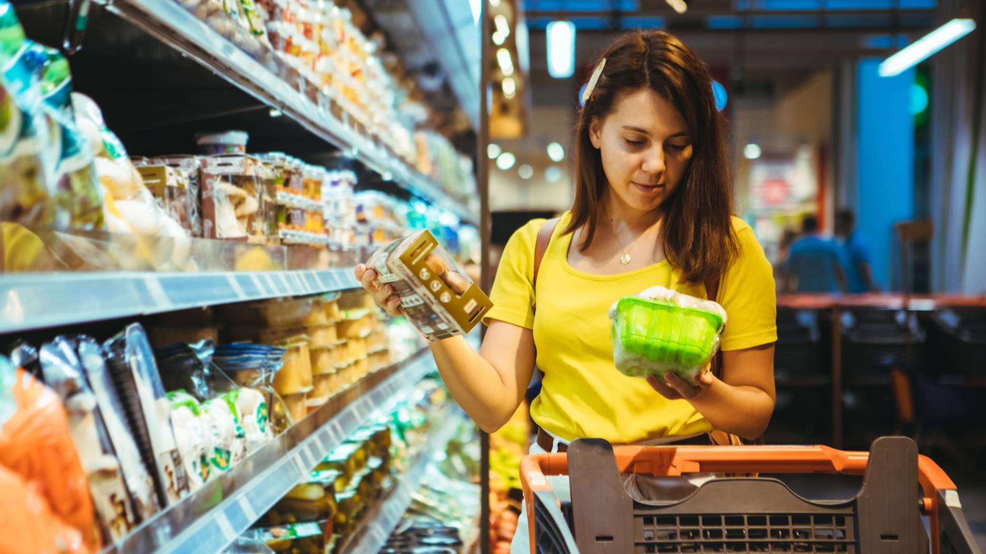 Eine Frau im gelben T-Shirt steht mit einem Einkaufswagen vorm Kühlregal. Sie schaut sich die Produkte genauer an.