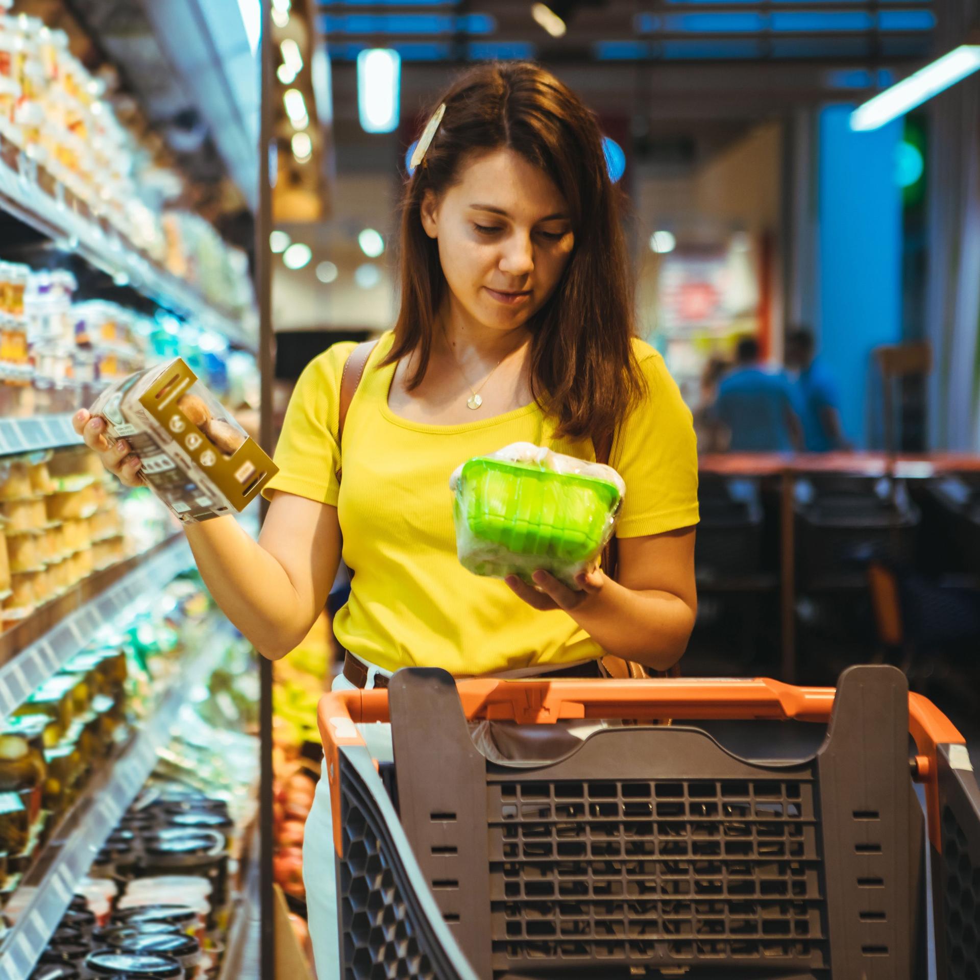 Eine Frau im gelben T-Shirt steht mit einem Einkaufswagen vorm Kühlregal. Sie schaut sich die Produkte genauer an.