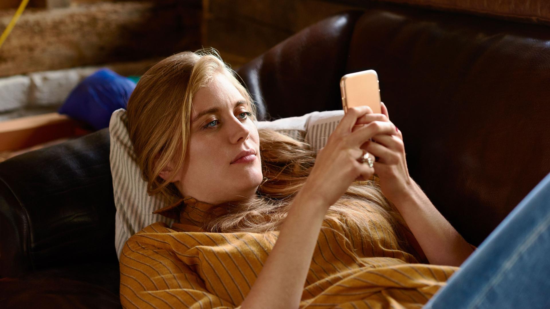 Eine blonde Frau liegt mit ihrem Handy in der Hand auf einem Sofa.