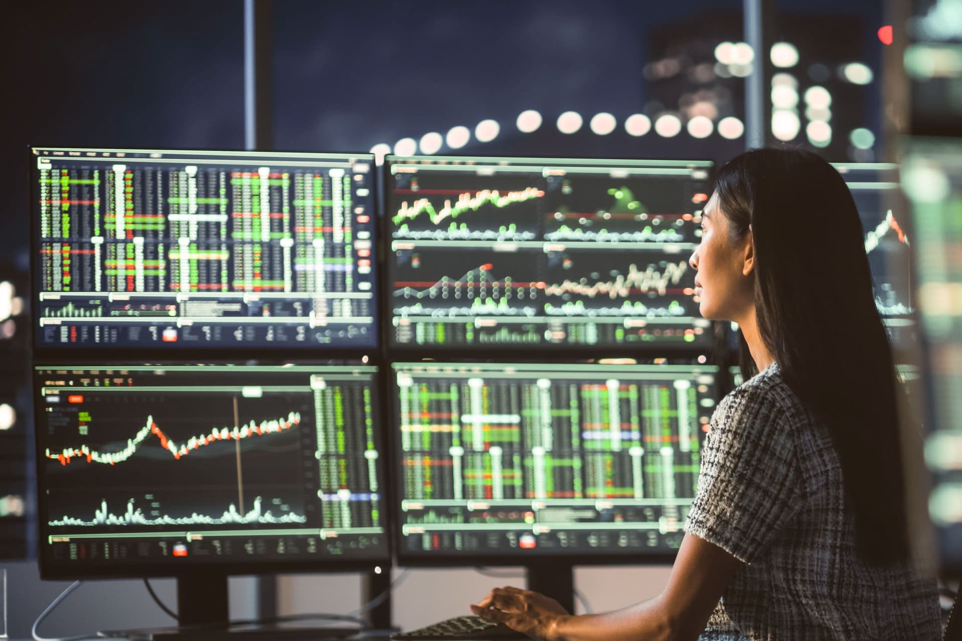 Eine Frau sitzt im Dunklen vor großen Monitoren, auf denen Börsenkurse abgebildet werden.
