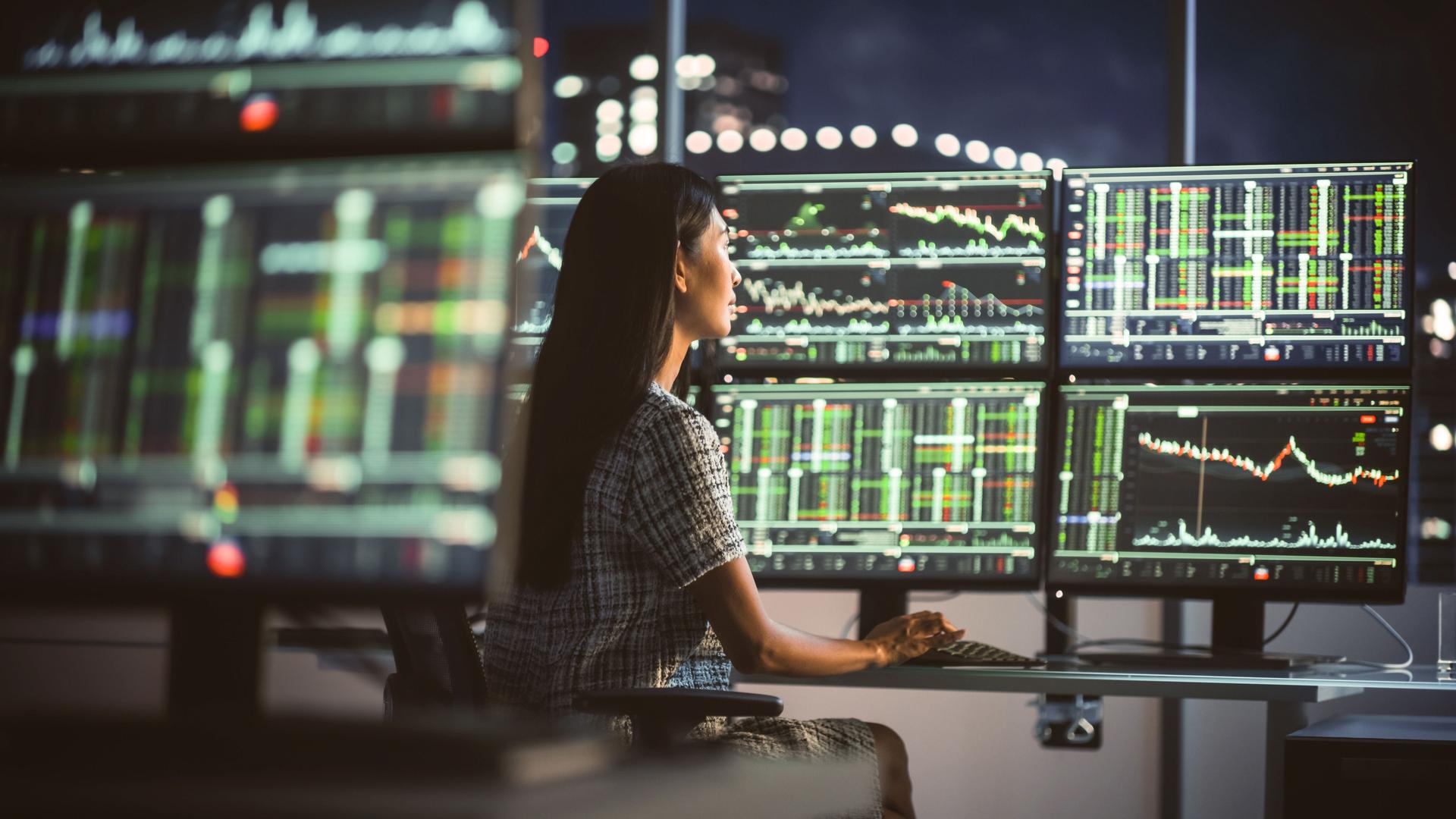 Eine Frau sitzt im Dunklen vor großen Monitoren, auf denen Börsenkurse abgebildet werden.