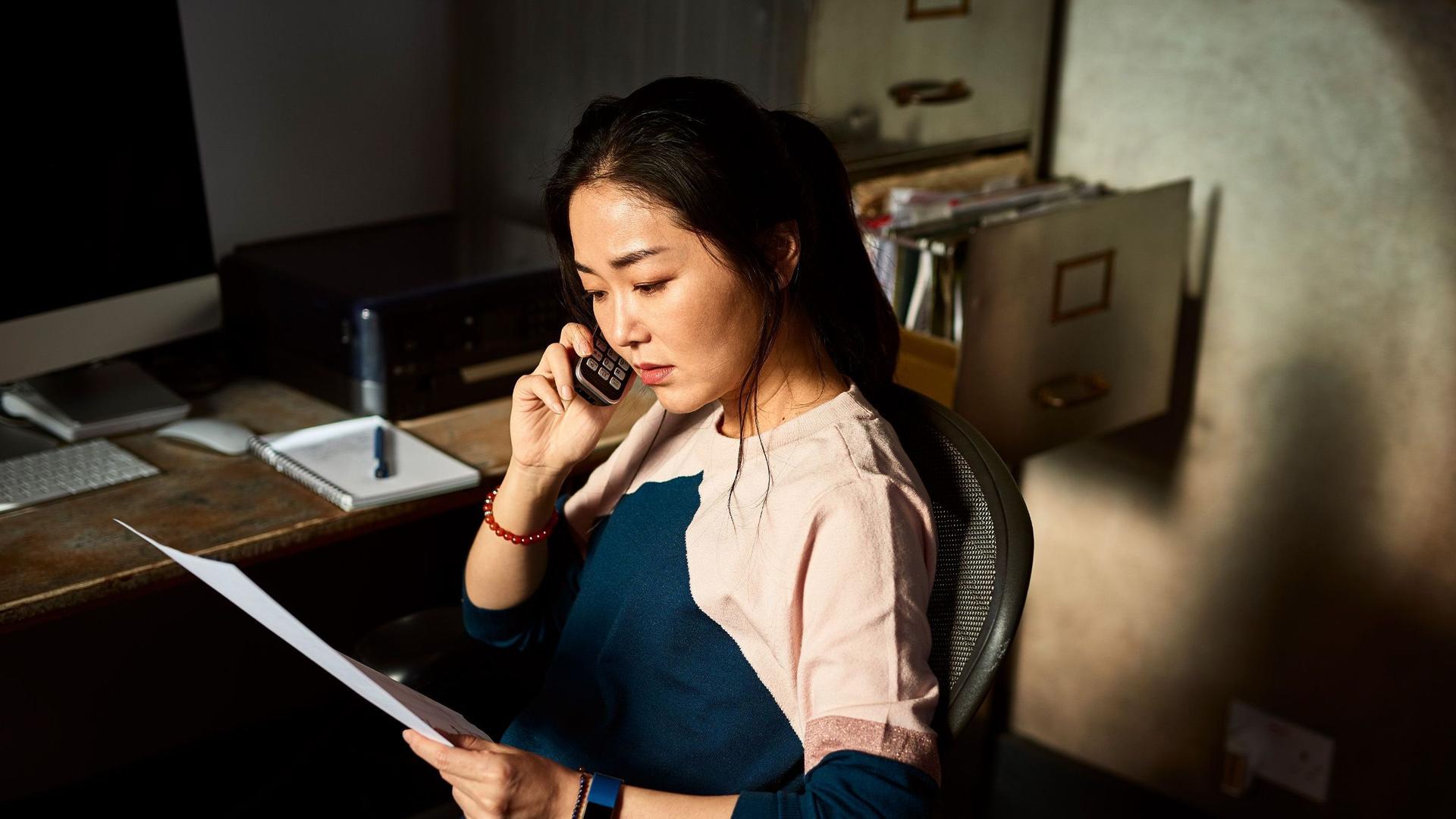 Eine Frau sitzt vor einem Schreibtisch schaut sorgenvoll auf eine Rechnung und telefoniert