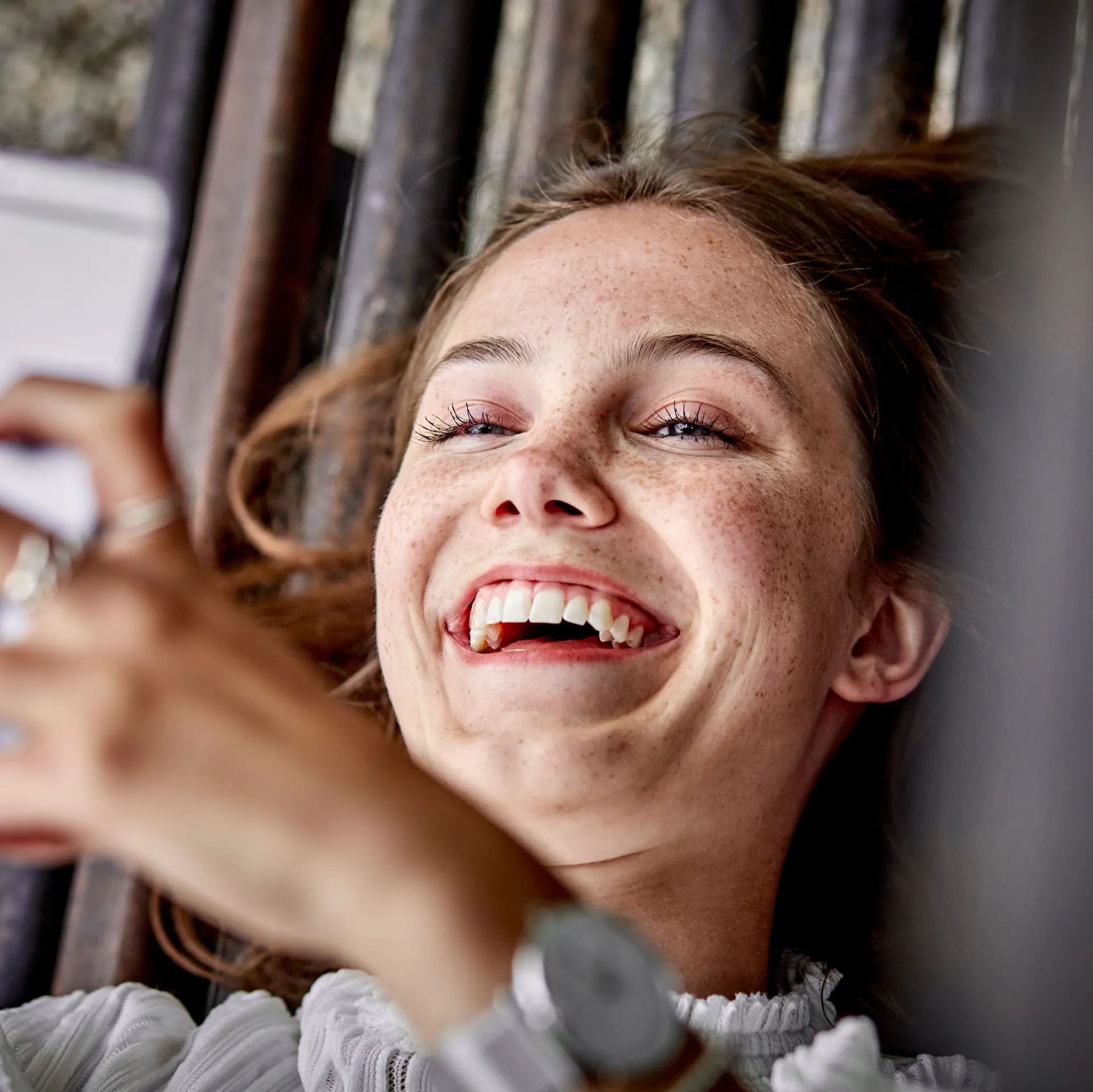 Eine junge Frau liegt auf einer Bank und hält ihr Handy Richtung Kamera. Sie lacht.