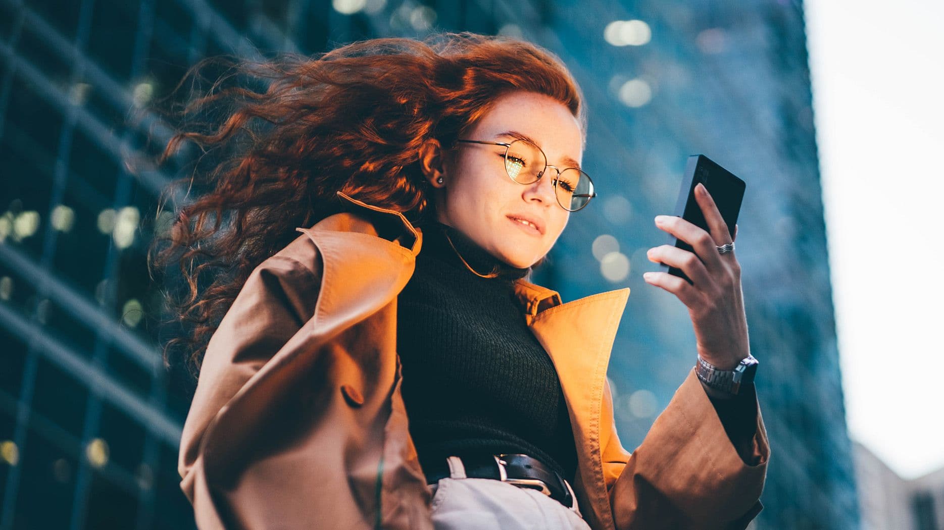 Junge Frau mit lockigem roten Haar und Brille schaut auf ihr Smartphone, im Hintergrund Glasfassaden eines Stadtgebäudes