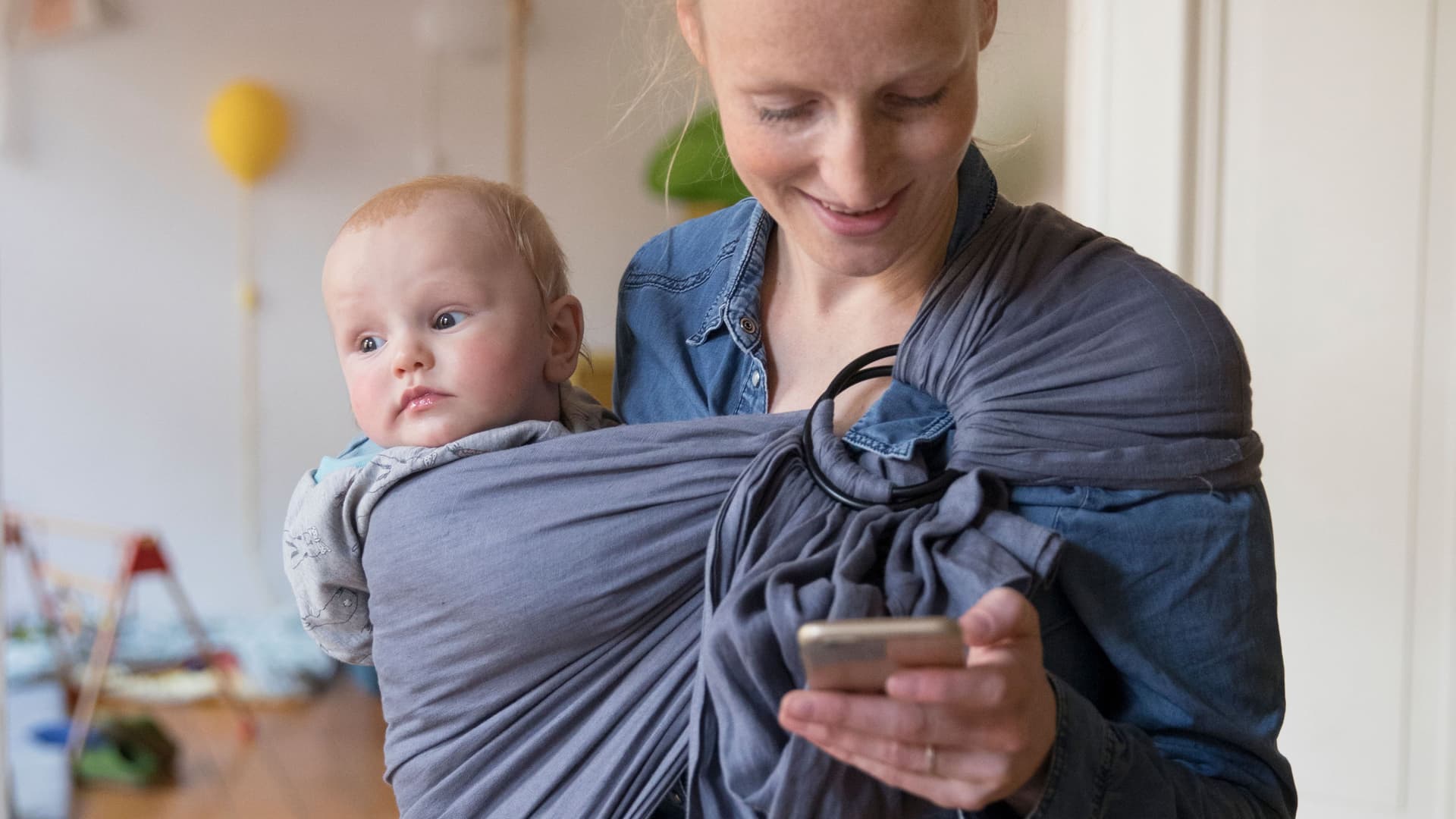 Eine junge Frau mit Kind im Tragetuch schaut auf ihr Handy. Sie lächelt.