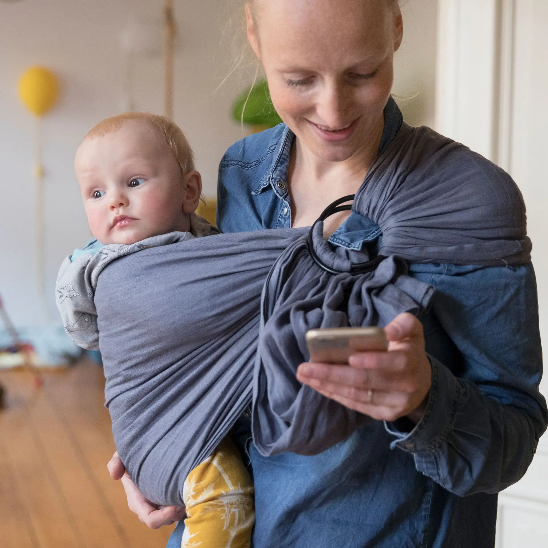 Eine junge Frau mit Kind im Tragetuch schaut auf ihr Handy. Sie lächelt.