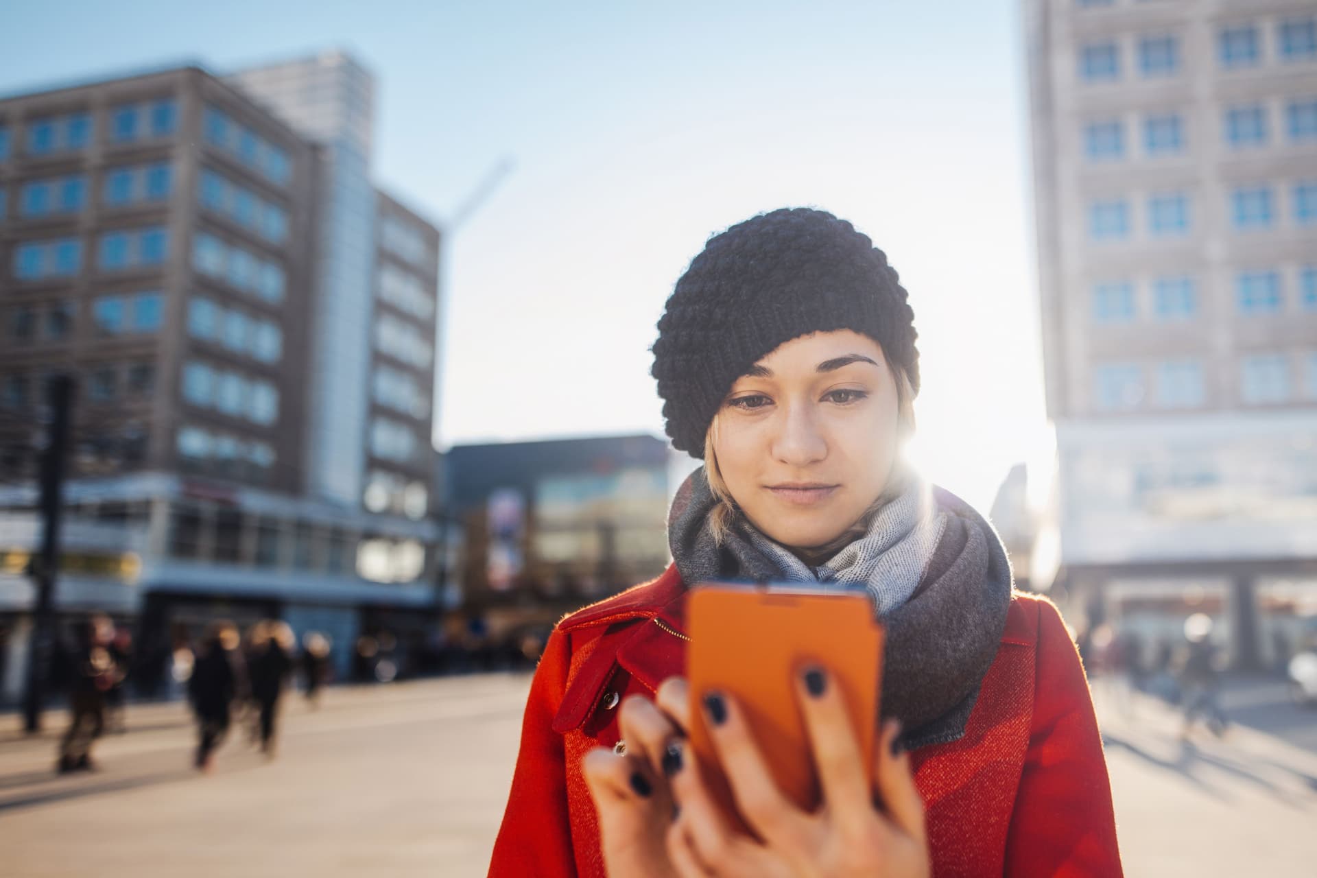 Eine junge Frau steht mit Mütze auf dem Kopf und einem Smartphone in der Hand steht auf dem Alexanderplatz mit der Sonne im Rücken.