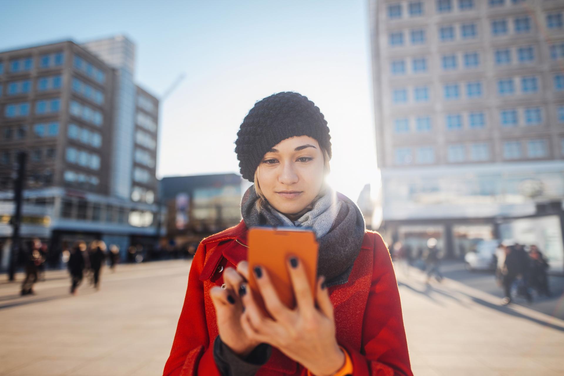 Eine junge Frau steht mit Mütze auf dem Kopf und einem Smartphone in der Hand steht auf dem Alexanderplatz mit der Sonne im Rücken.