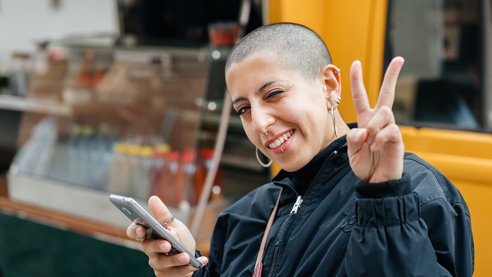 Eine junge Frau mit kurzrasierten Haaren, die ein Smartphone hält, lächelt in die Kamera und zeigt mit zwei Fingern das Victory-Zeichen.