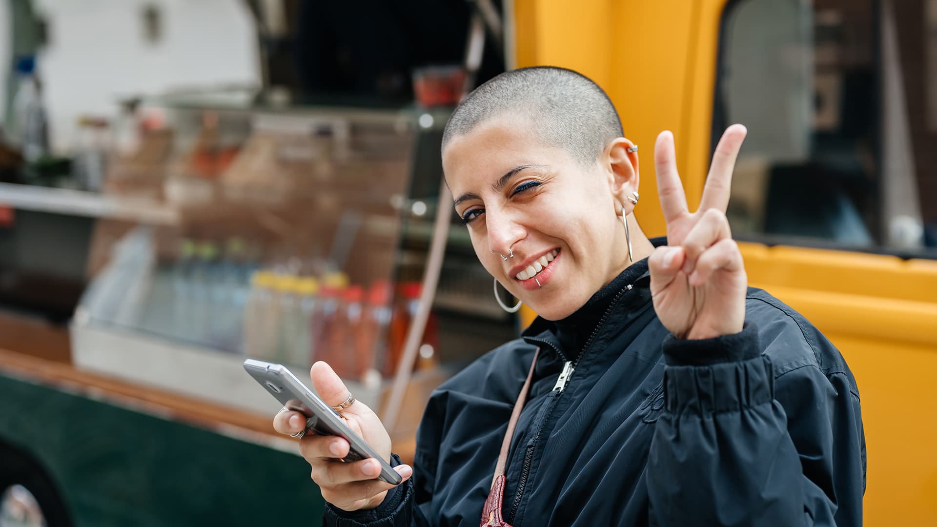 Eine junge Frau mit kurzrasierten Haaren, die ein Smartphone hält, lächelt in die Kamera und zeigt mit zwei Fingern das Victory-Zeichen.